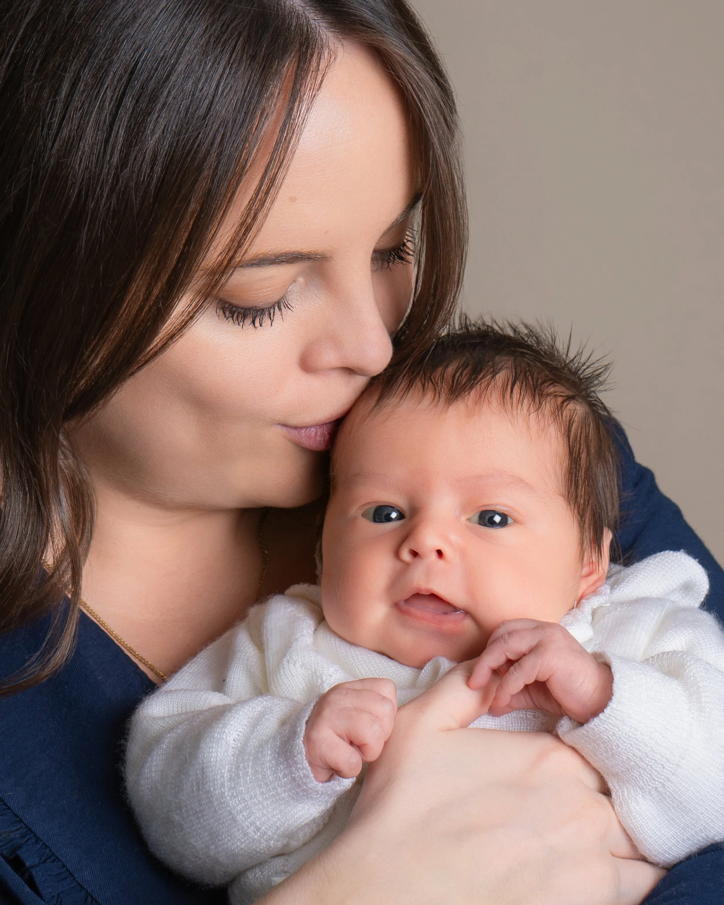A woman with long brown hair holding and kissing a baby with blue eyes, light skin, and dark hair wearing a cream sweater.