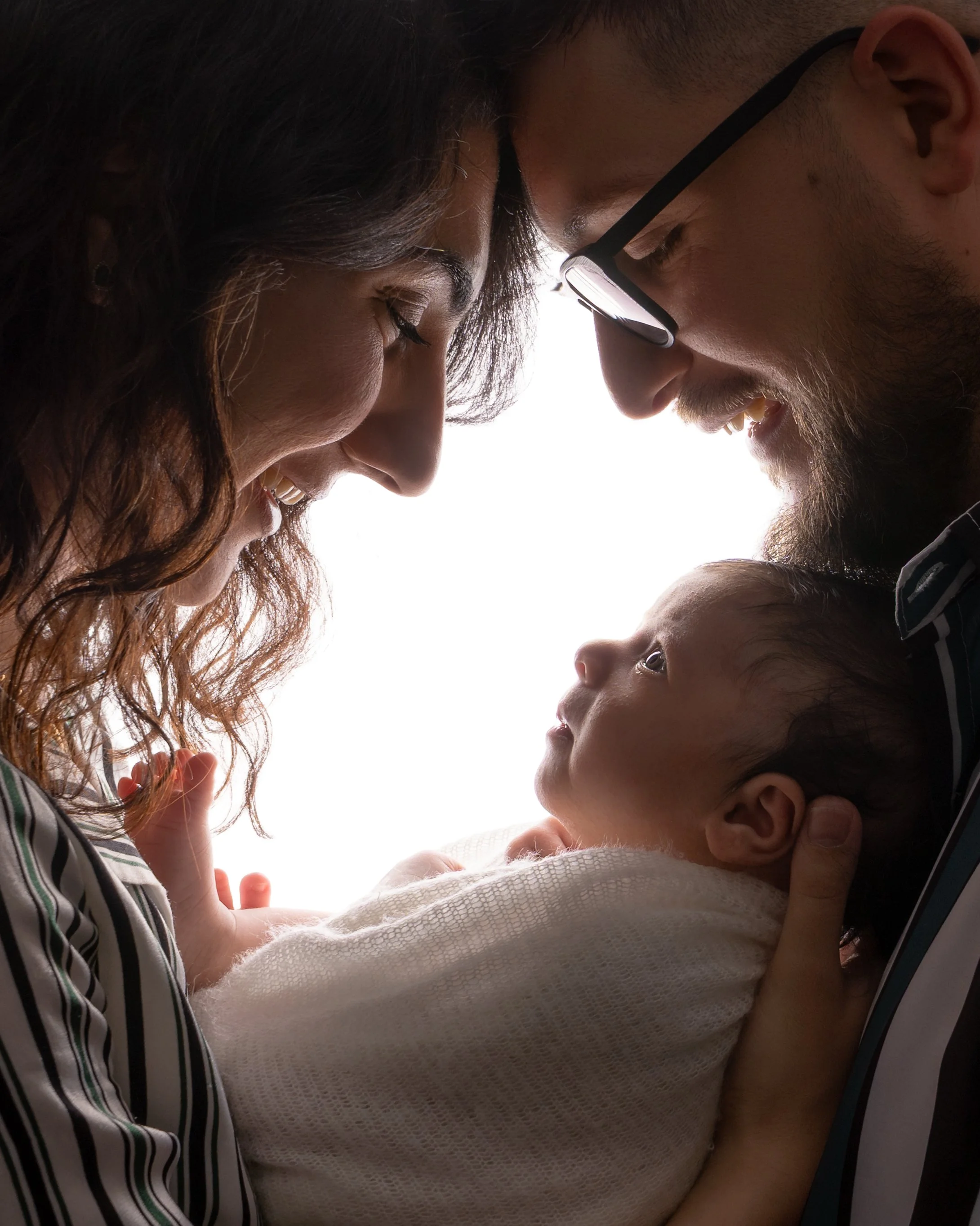 A woman and a man with glasses, facing each other closely, sharing a moment of affection with their foreheads touching and eyes closed, and a young girl looking up at them with a gentle expression.