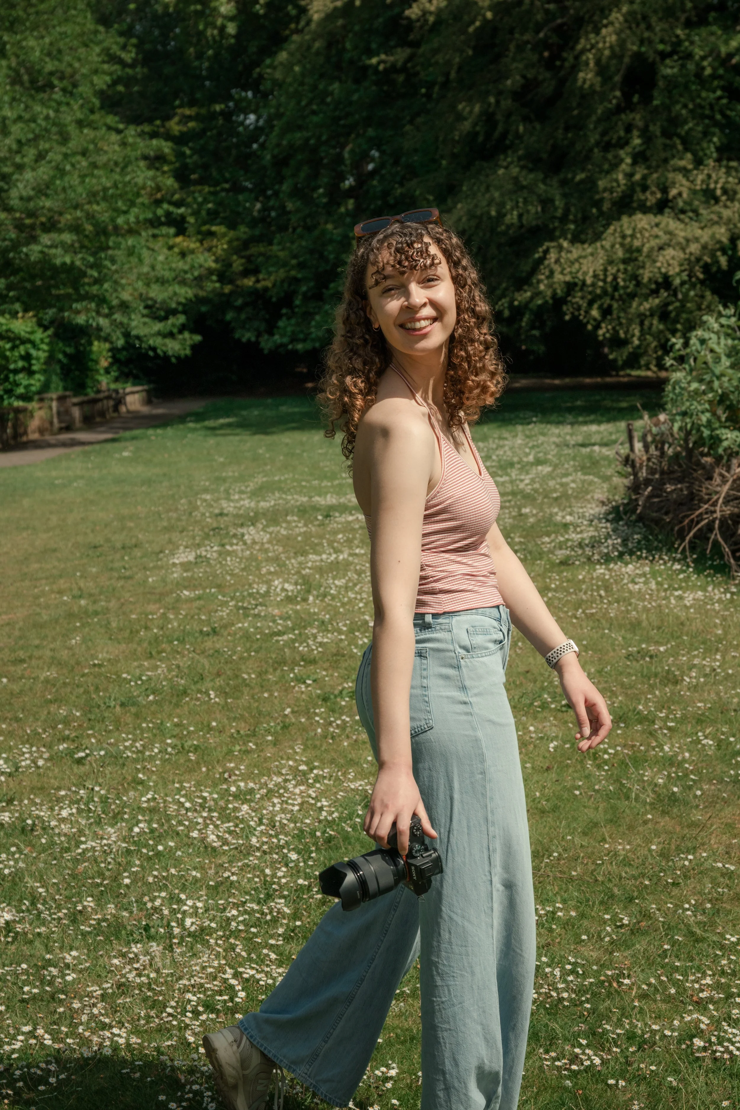 A young woman with curly hair in a striped tank top and wide-leg jeans stands outdoors in a grassy park, smiling and holding a camera, with sunglasses on her head.