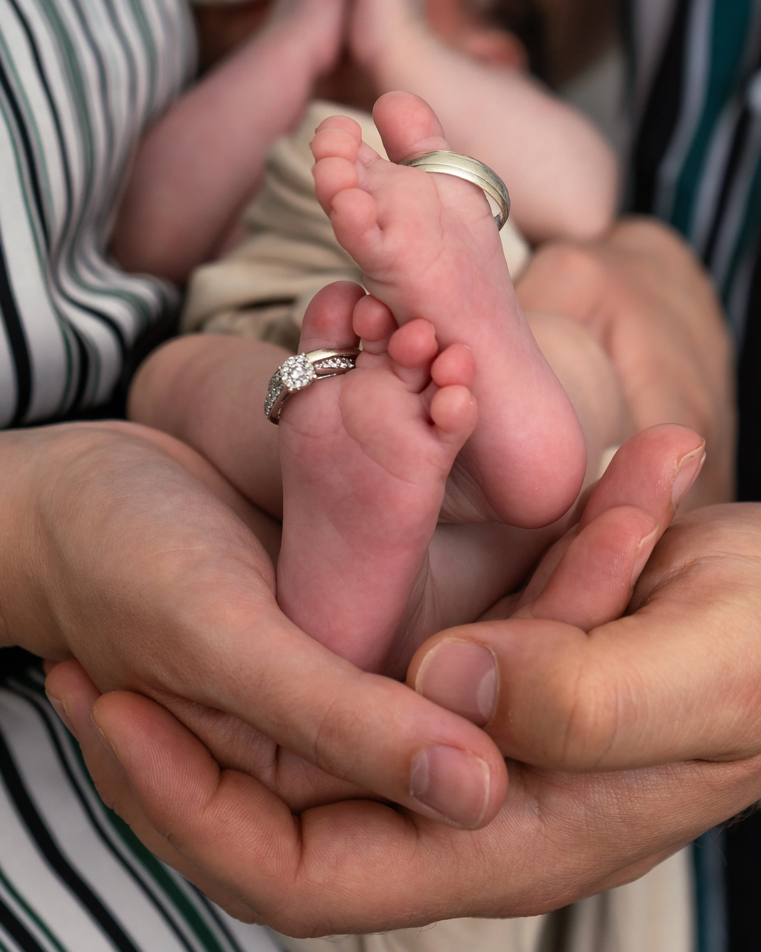 Close-up of a parent's hand holding a baby's foot, with a wedding ring on the parent's finger and a ring with diamonds on the baby's toe.