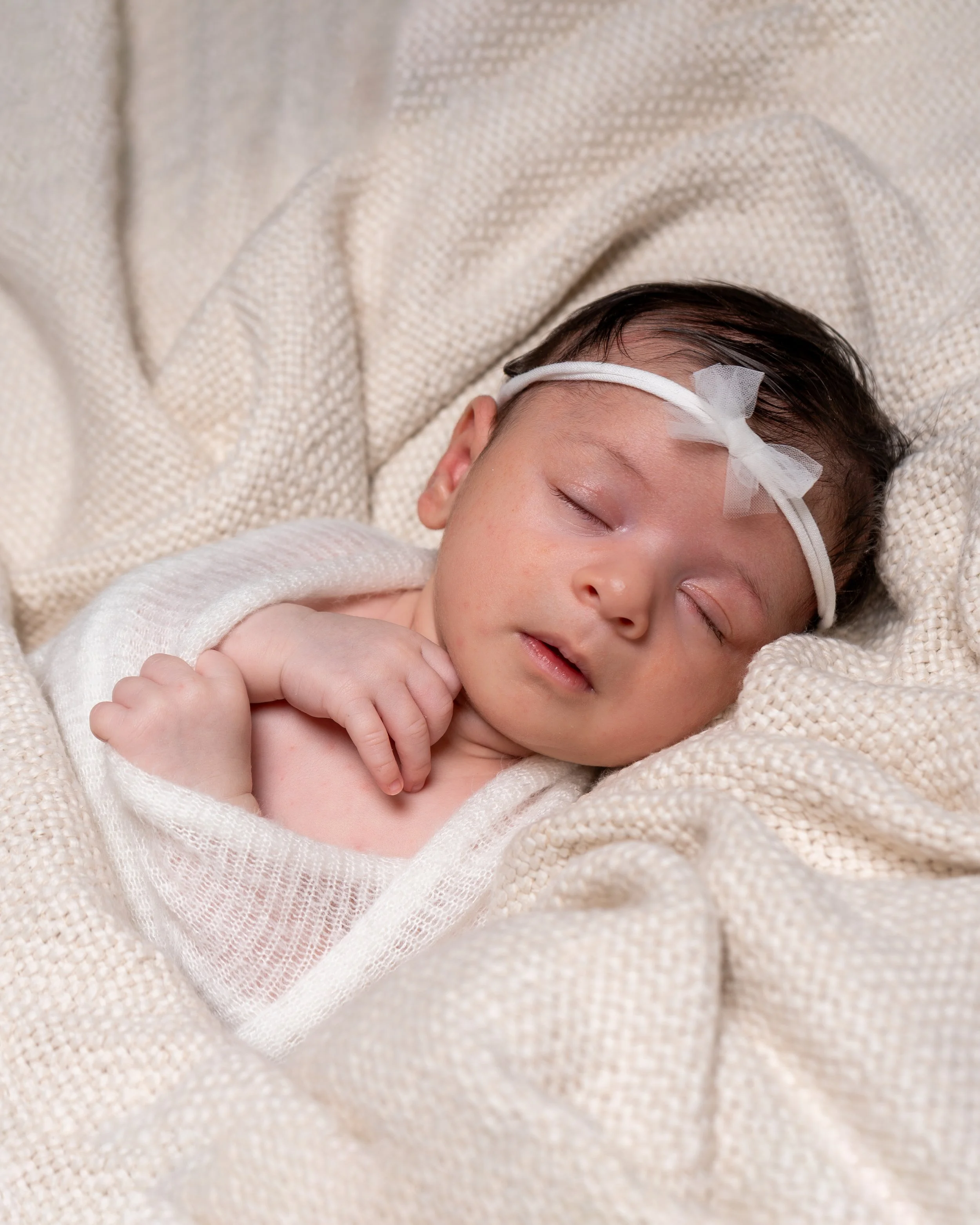 Close-up of a sleeping baby with dark hair, wearing a white headband with a bow, wrapped in a cream-colored blanket.