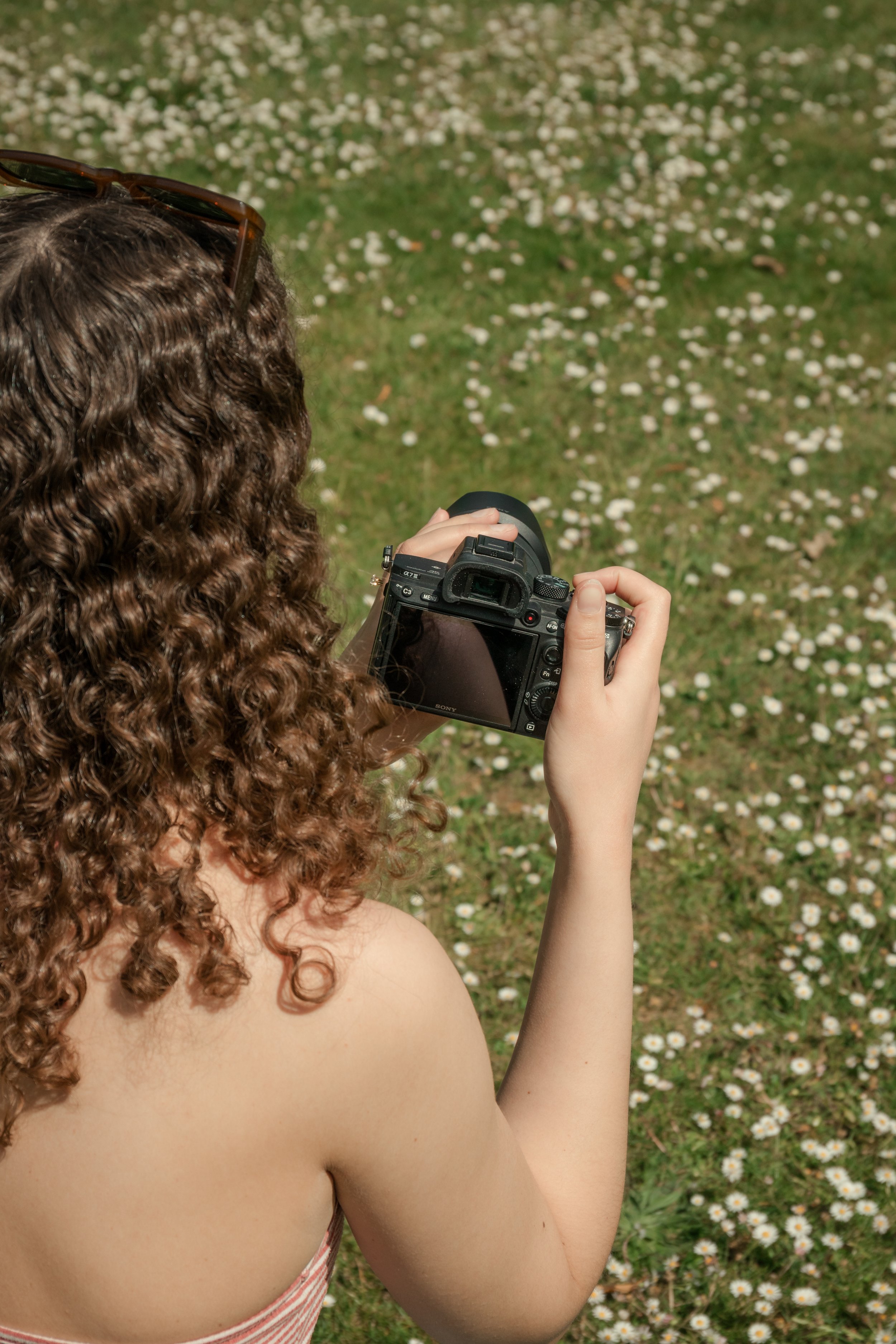 A woman with long curly hair is holding a camera and taking a picture outdoors. She is standing in a garden with small white flowers on the grass.