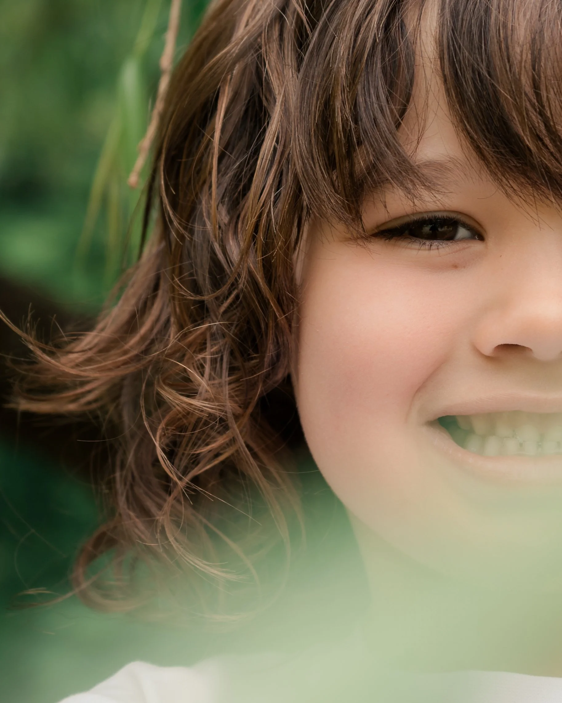 A close-up of a child's face with brown, wavy hair and dark eyes, smiling outdoors with greenery in the background.