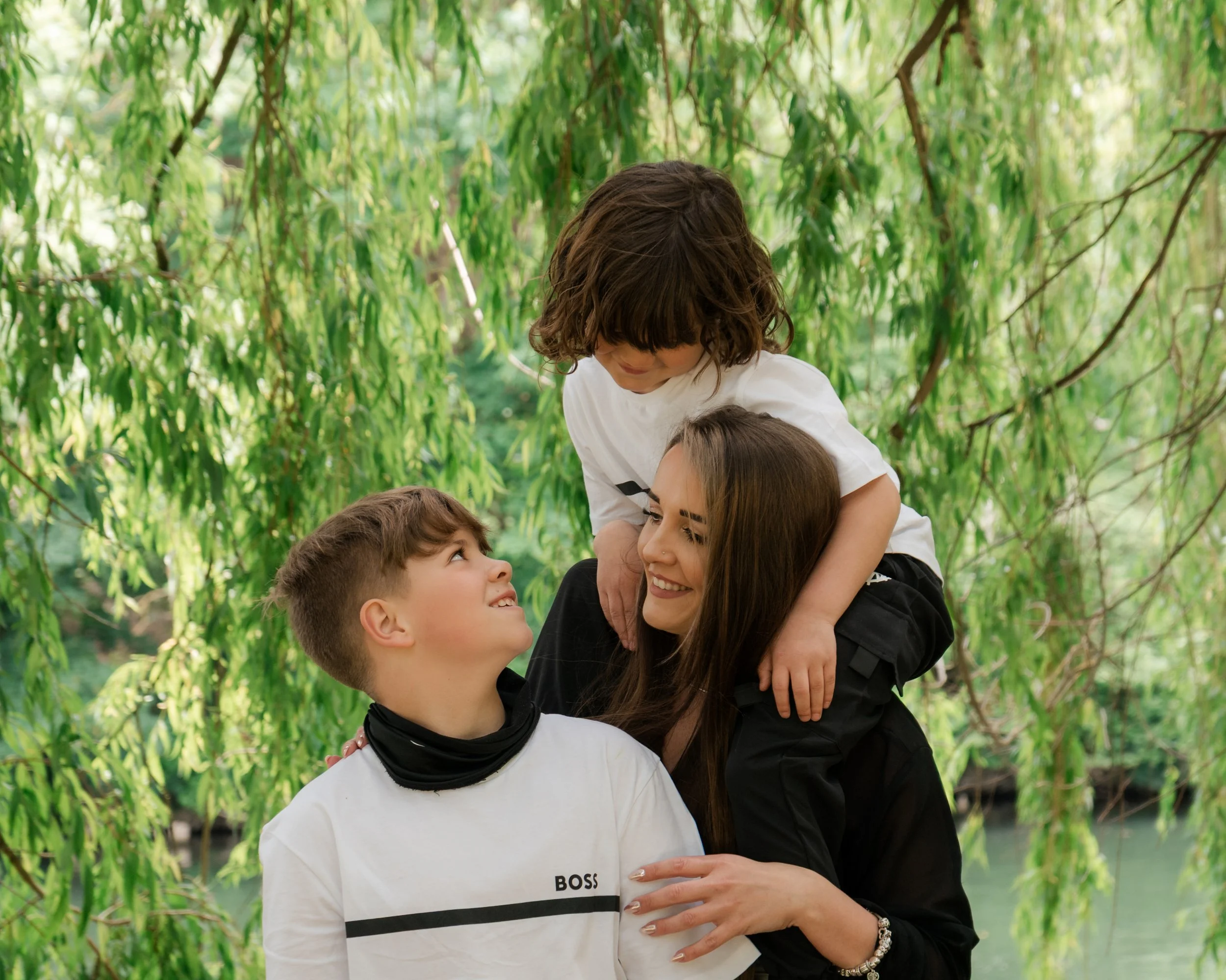 A woman with long brown hair and a black top is embracing a young boy with short brown hair, who is looking up at her. Two other children are in the foreground, one with short hair and the other with longer light brown hair, looking at the woman. They are outdoors under green, leafy trees.