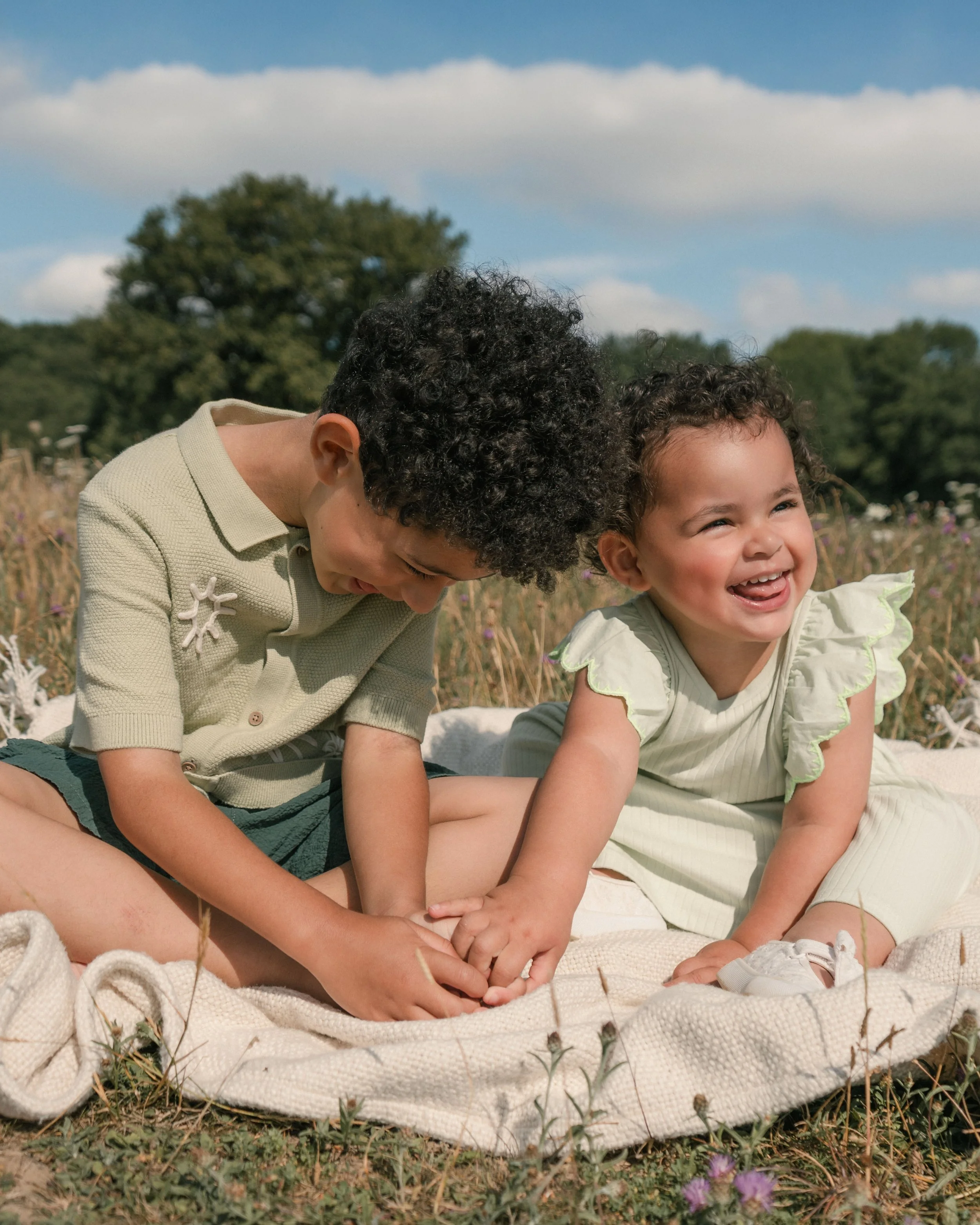 Two children, a boy and a girl, sitting outdoors on a blanket, smiling and laughing, surrounded by grass and wildflowers, with trees and a partly cloudy sky in the background.