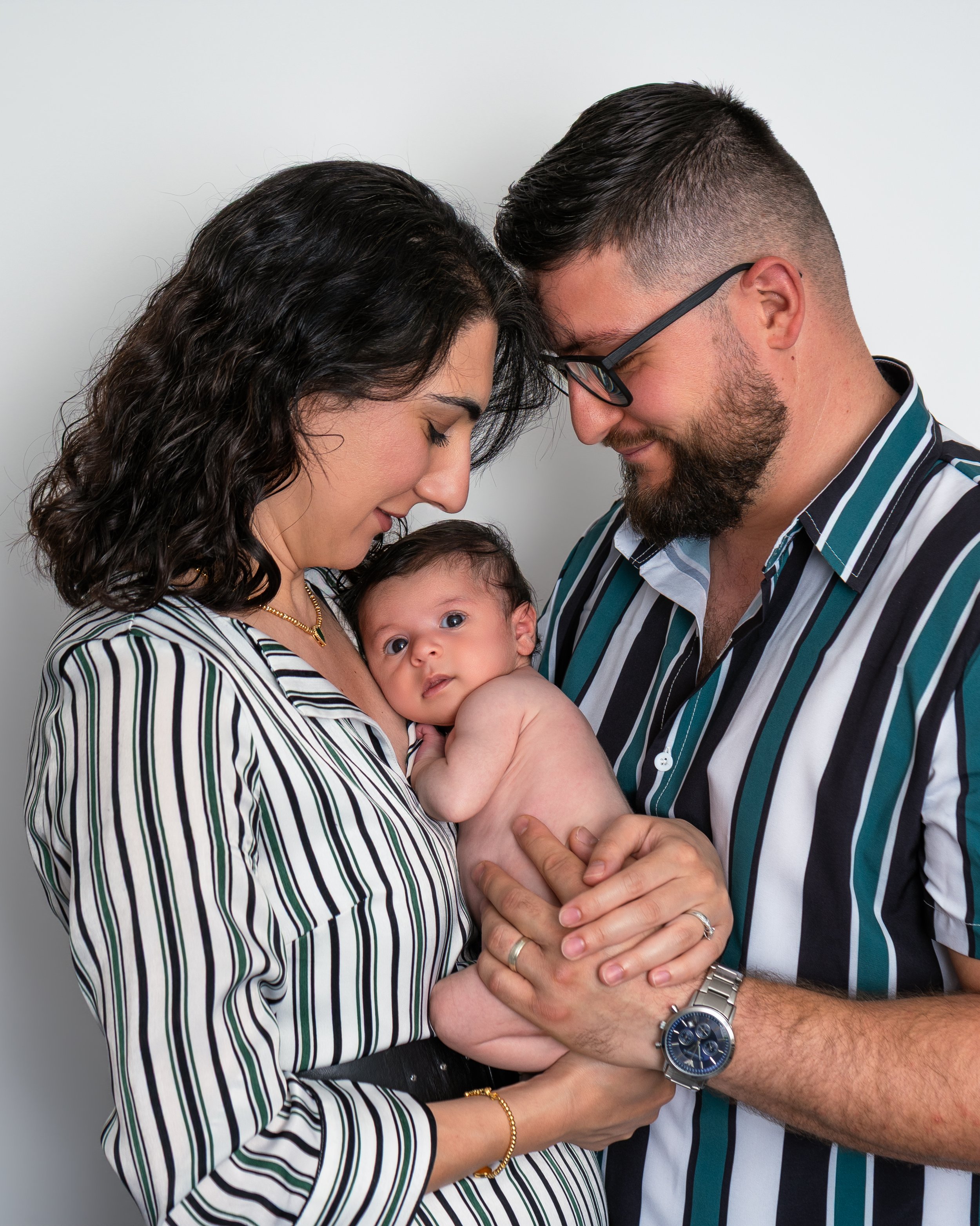 A family of three, including a man, a woman, and a baby, shares a tender moment with their foreheads touching. The woman and the man are holding the baby between them, and they are all smiling softly. The woman has wavy dark hair and is wearing a striped shirt along with a gold bracelet. The man has a beard, glasses, and is wearing a striped shirt, along with a silver watch. The baby is looking directly at the camera and appears to be a few months old.