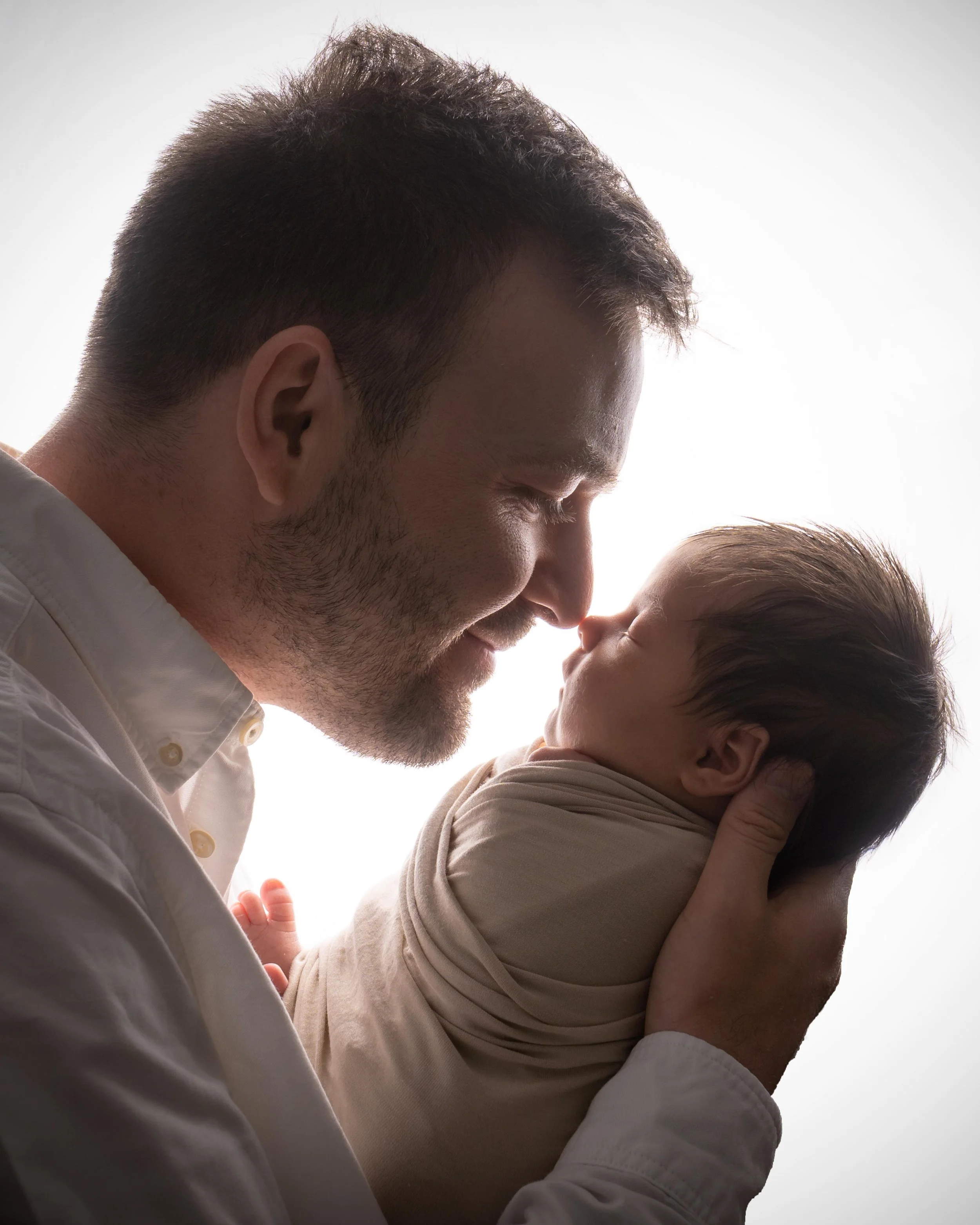 A man with short dark hair and light facial hair holds a young child close to his face, both with eyes closed, touching foreheads, backlit with bright light creating a silhouette effect.