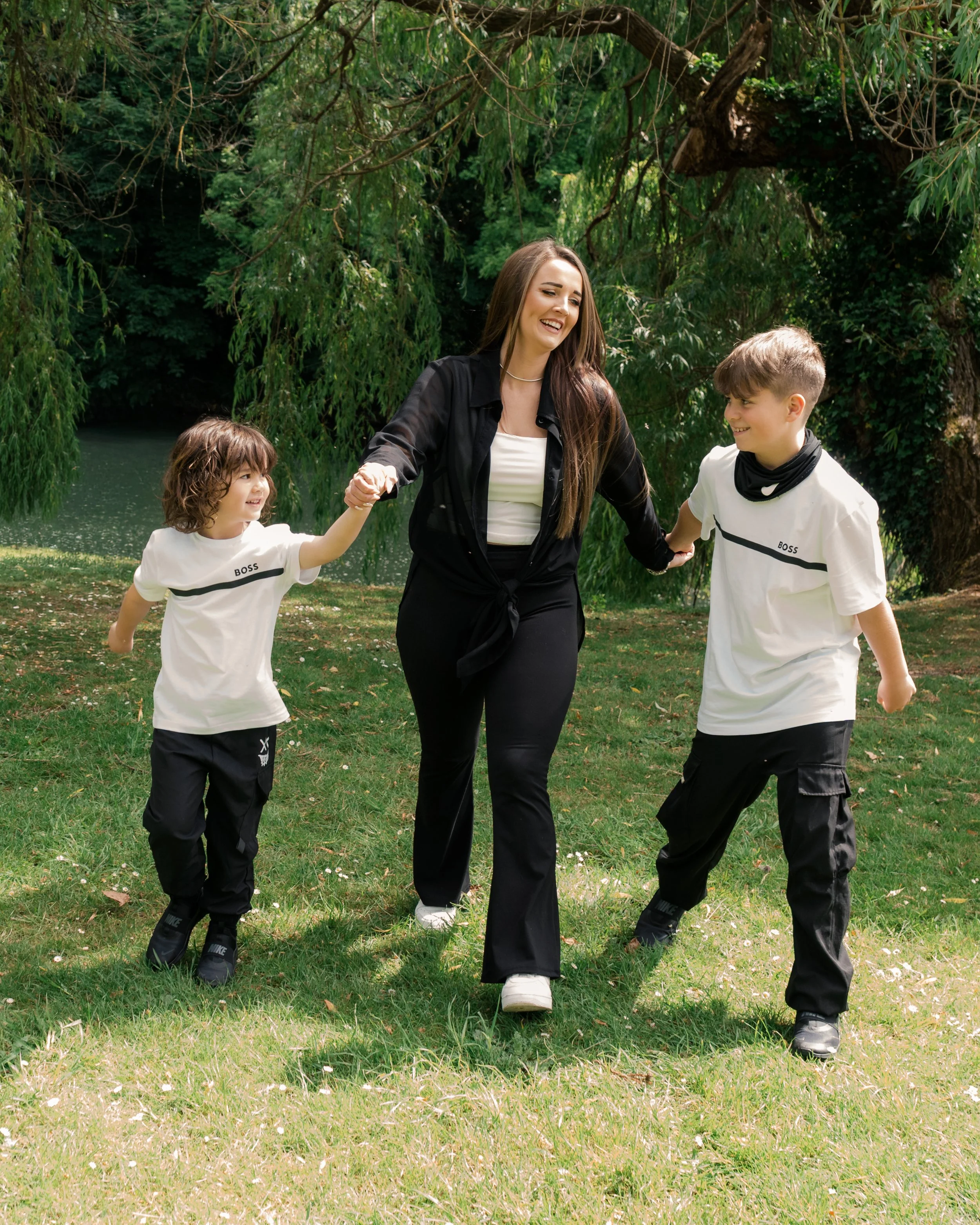 A woman with long brown hair smiling and holding hands with two children, a girl and a boy, as they walk through a grassy area near trees and a pond on a sunny day.