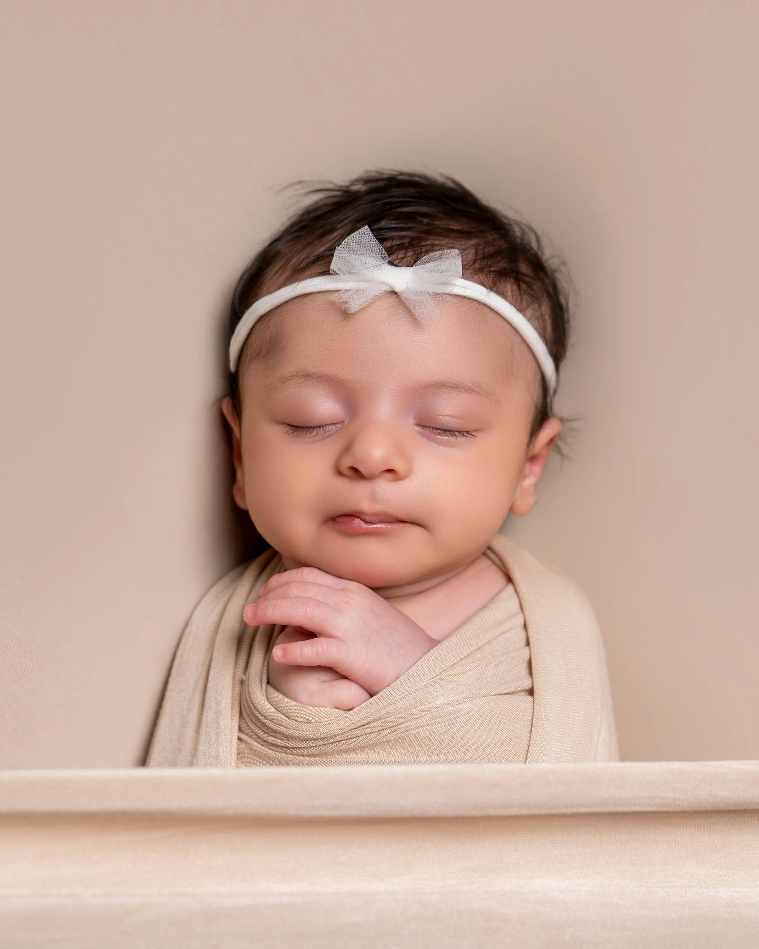 A baby girl with closed eyes and a peaceful expression, wearing a white headband with a bow, lying in a beige box or cradle with a neutral background.