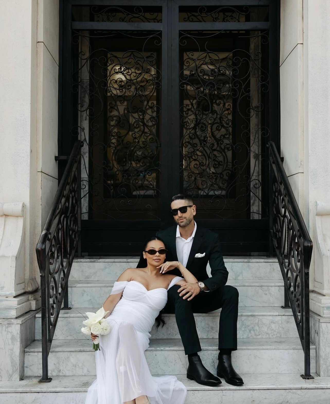 A man and woman sitting on the steps of a building with ornate black wrought iron doors behind them. The woman is wearing a white off-the-shoulder dress and holding a white bouquet. The man is in a black suit, white shirt, and sunglasses.