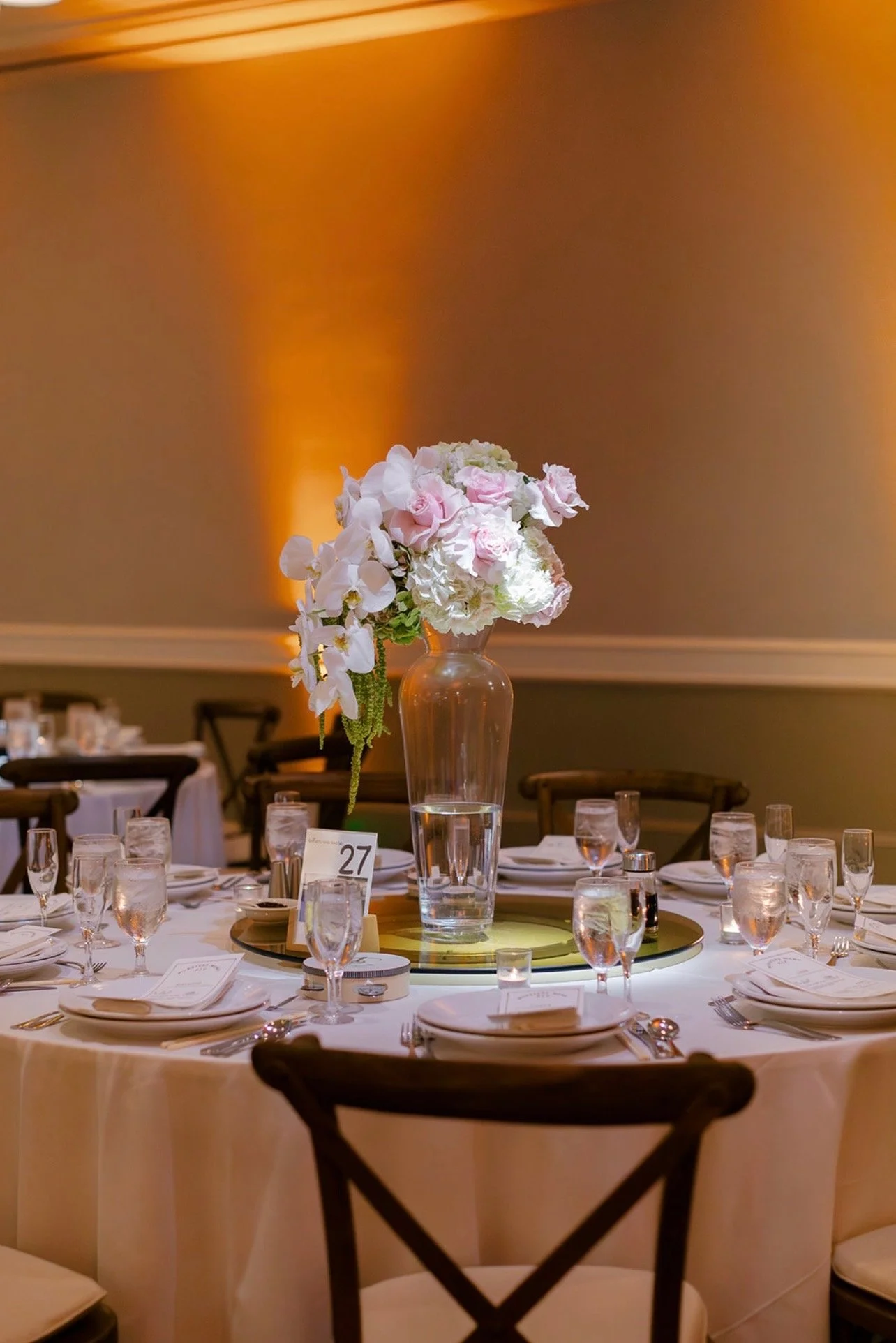 Elegant banquet table setup with a large flower arrangement centerpiece in a transparent vase, surrounded by glasses, plates, and utensils, with warm lighting in the background.