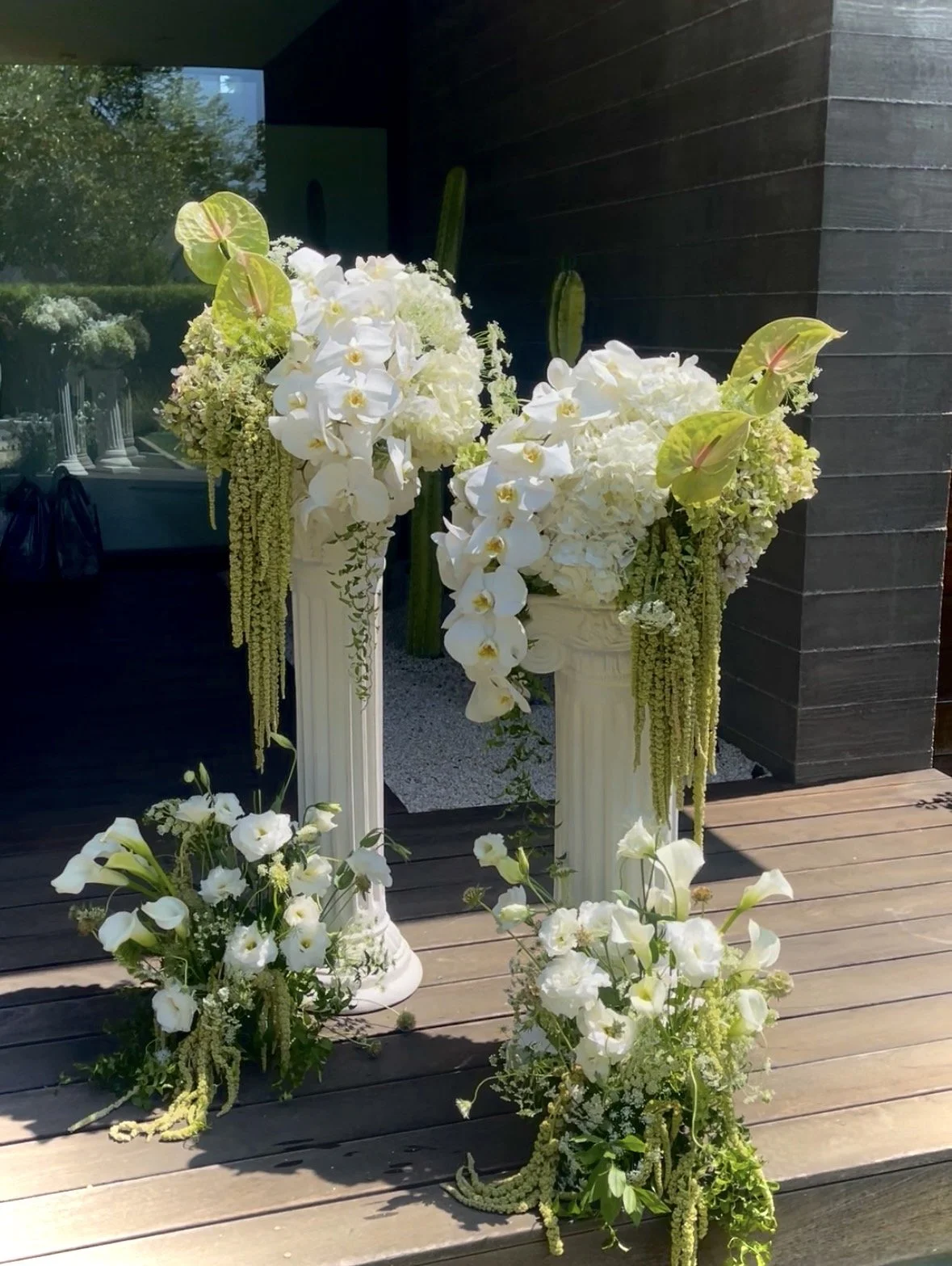 Elegant floral arrangement with white orchids, hydrangeas, calla lilies, and hanging amaranthus on tall, white Corinthian-style pedestals, set against a dark wall and outdoor patio.