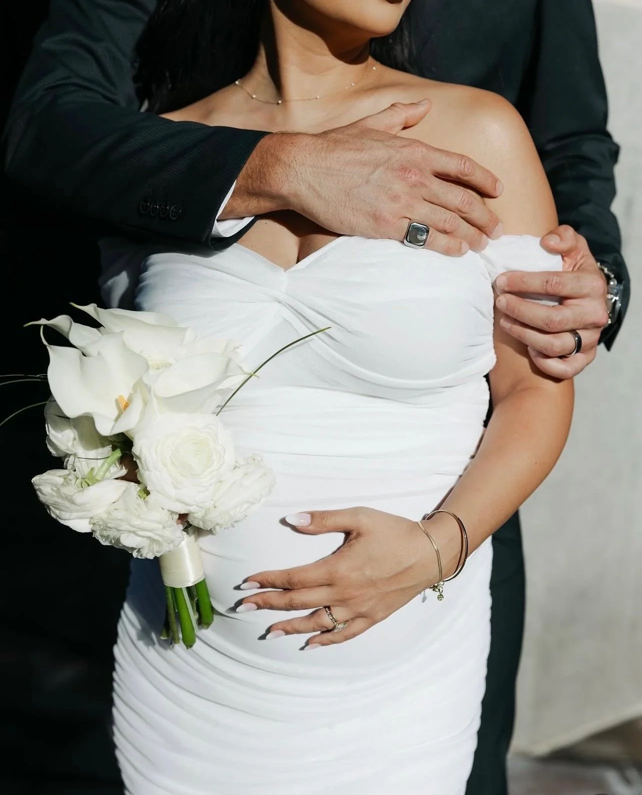 Close-up of a woman in a white wedding dress holding a bouquet of white flowers, with a man in a black suit behind her, both embracing.