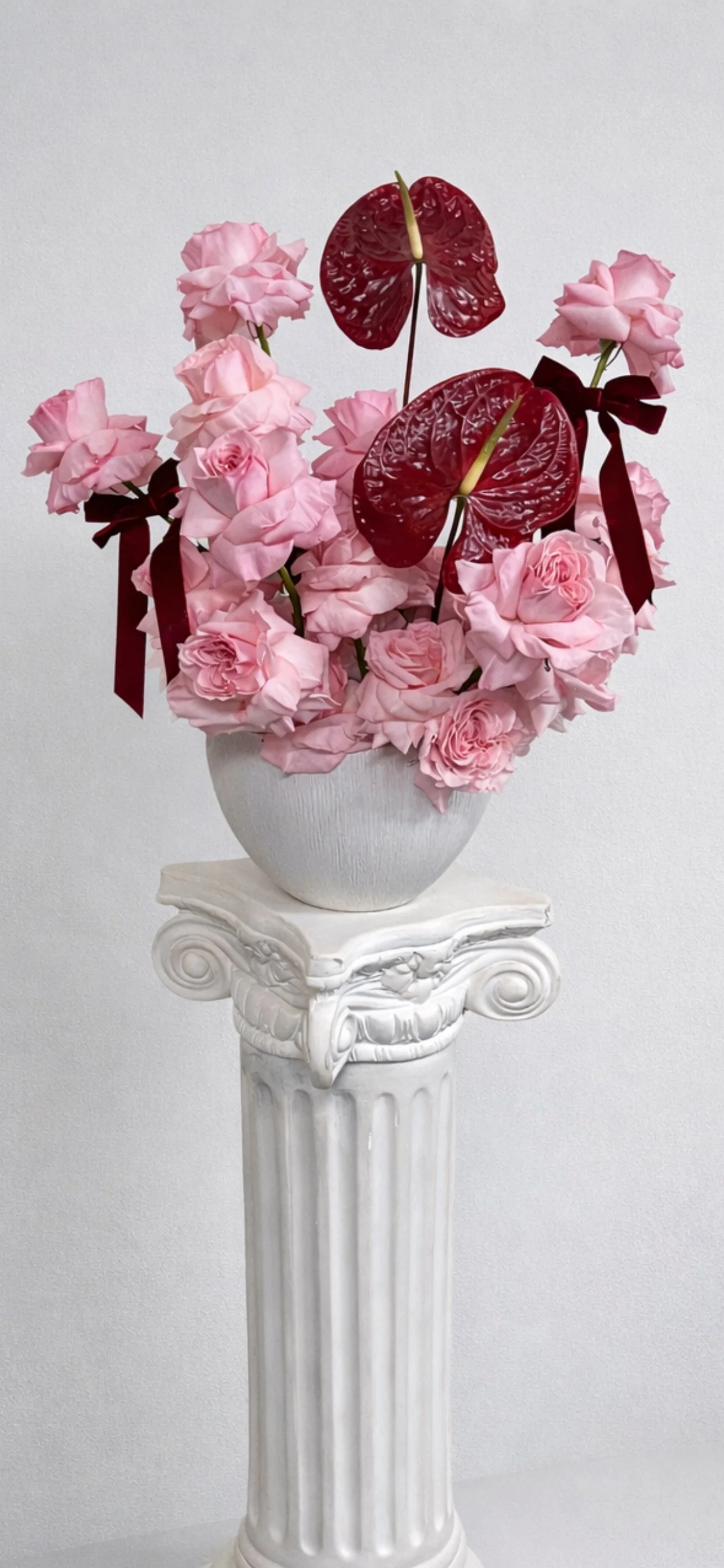 Artificial flower arrangement with pink roses and red anthuriums in a white vase on a decorative white pedestal.