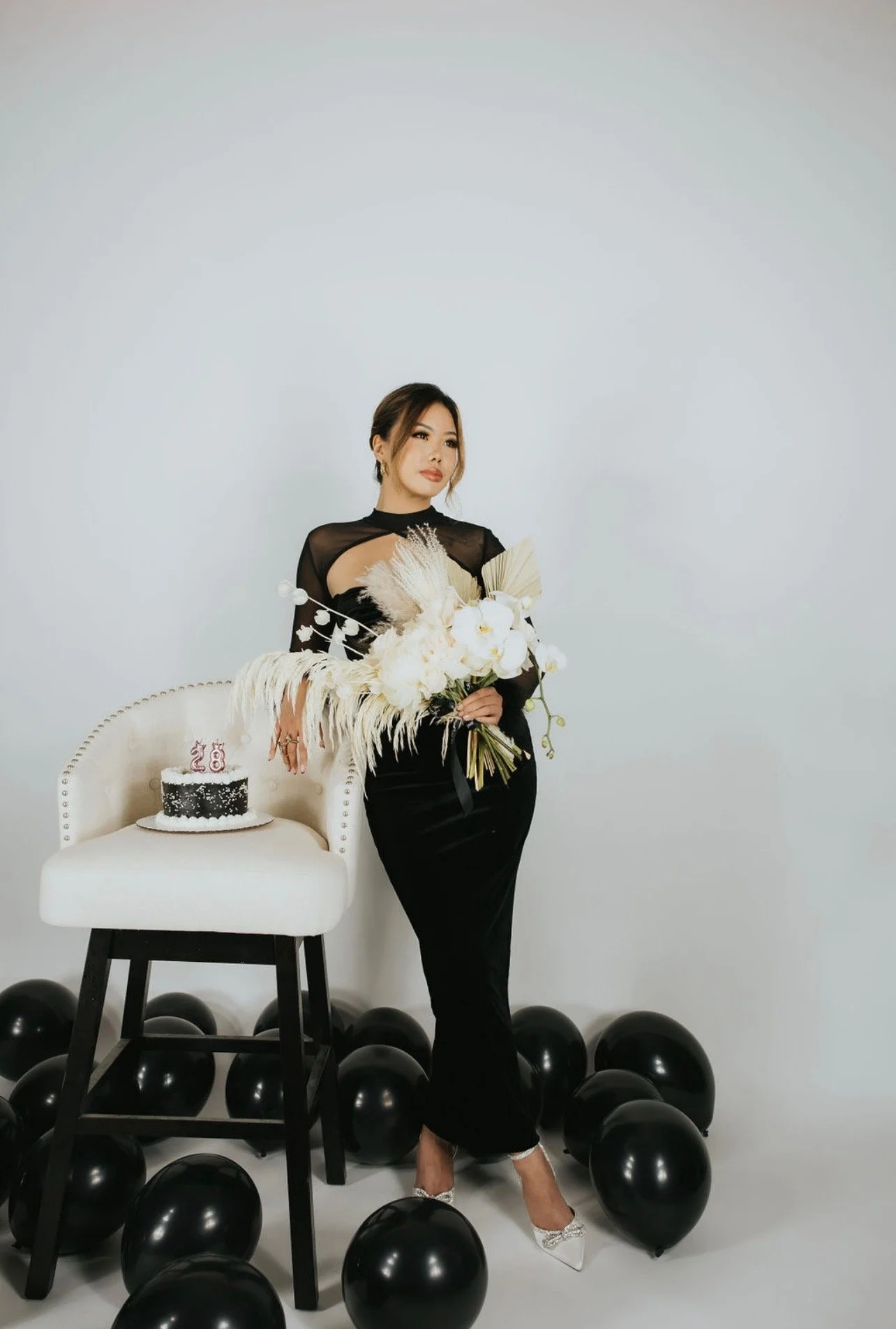 A woman in a black dress holding a bouquet of white flowers at a celebration, with black balloons and a small black and white cake with candles that read '28' on a white chair next to her.