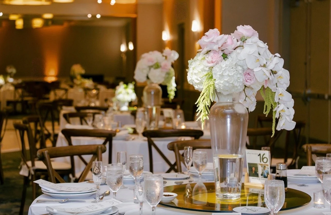 Elegant banquet hall with round tables decorated with pink and white flower centerpieces, glassware, and table settings.