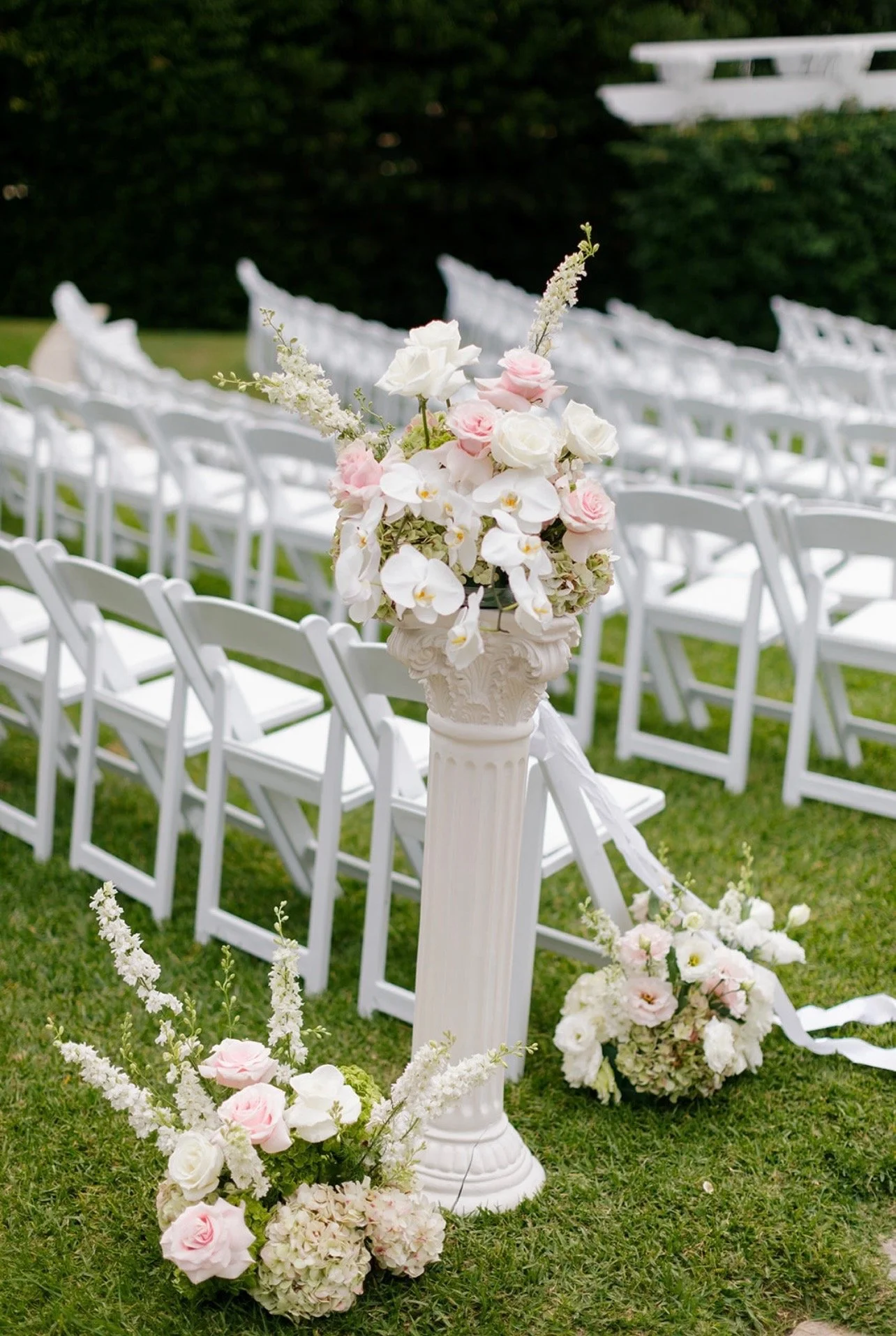 White chairs arranged in rows outdoors, decorated with floral arrangements of white and pink roses, hydrangeas, and other flowers, for a wedding ceremony.