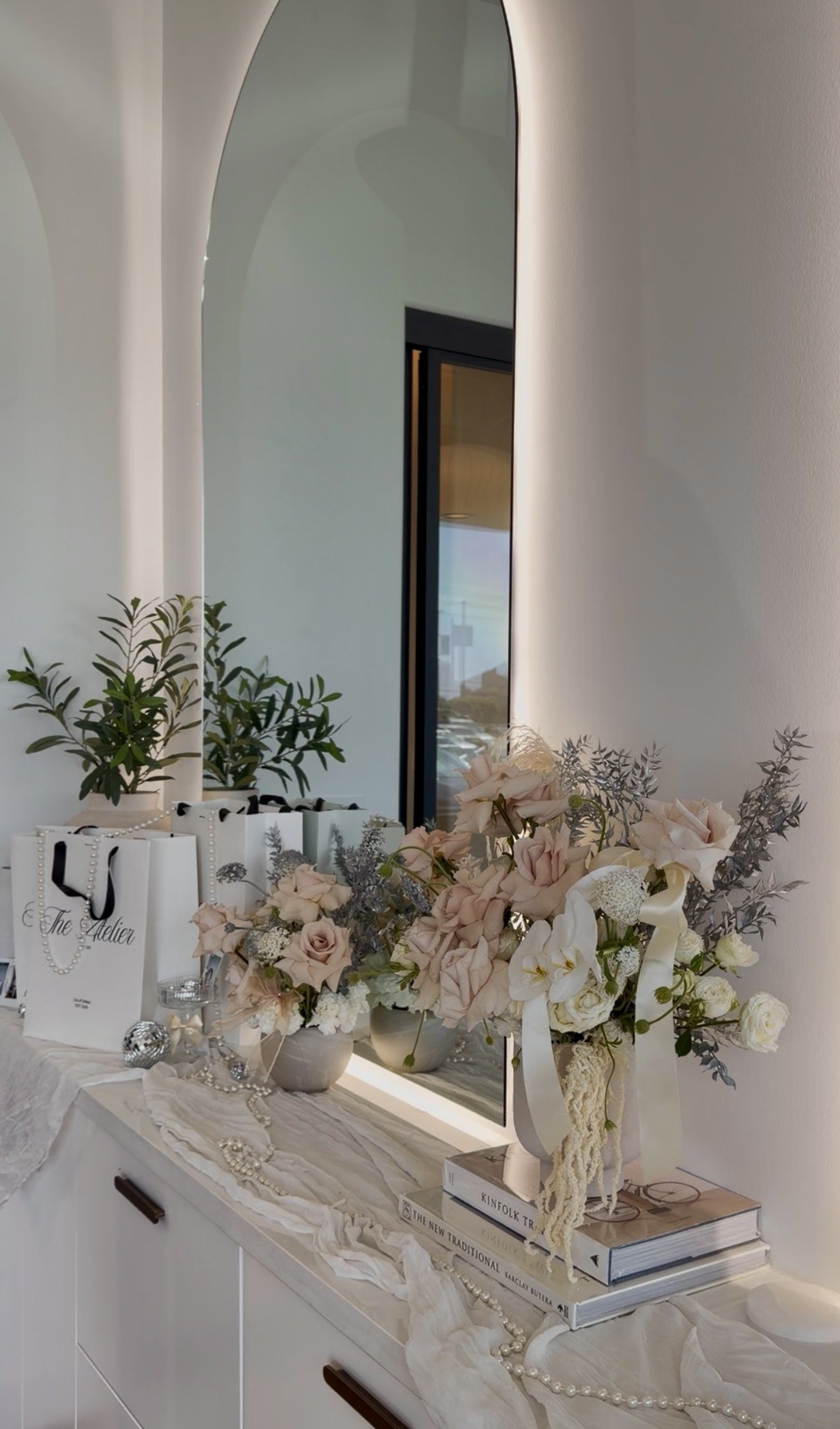A dresser decorated with a large bouquet of pale pink, white, and lavender flowers, some white gift bags with black handles, a string of pearls, and scattered jewelry, with a mirror on a white wall reflecting part of a room and a window.