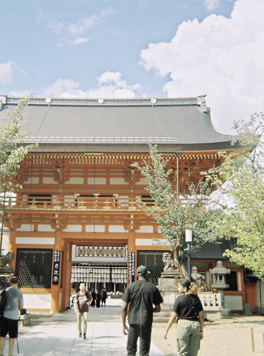 Une entrée d'un temple japonais traditionnel avec des personnes qui entrent et sortent, entouré d'arbres et de lanternes.