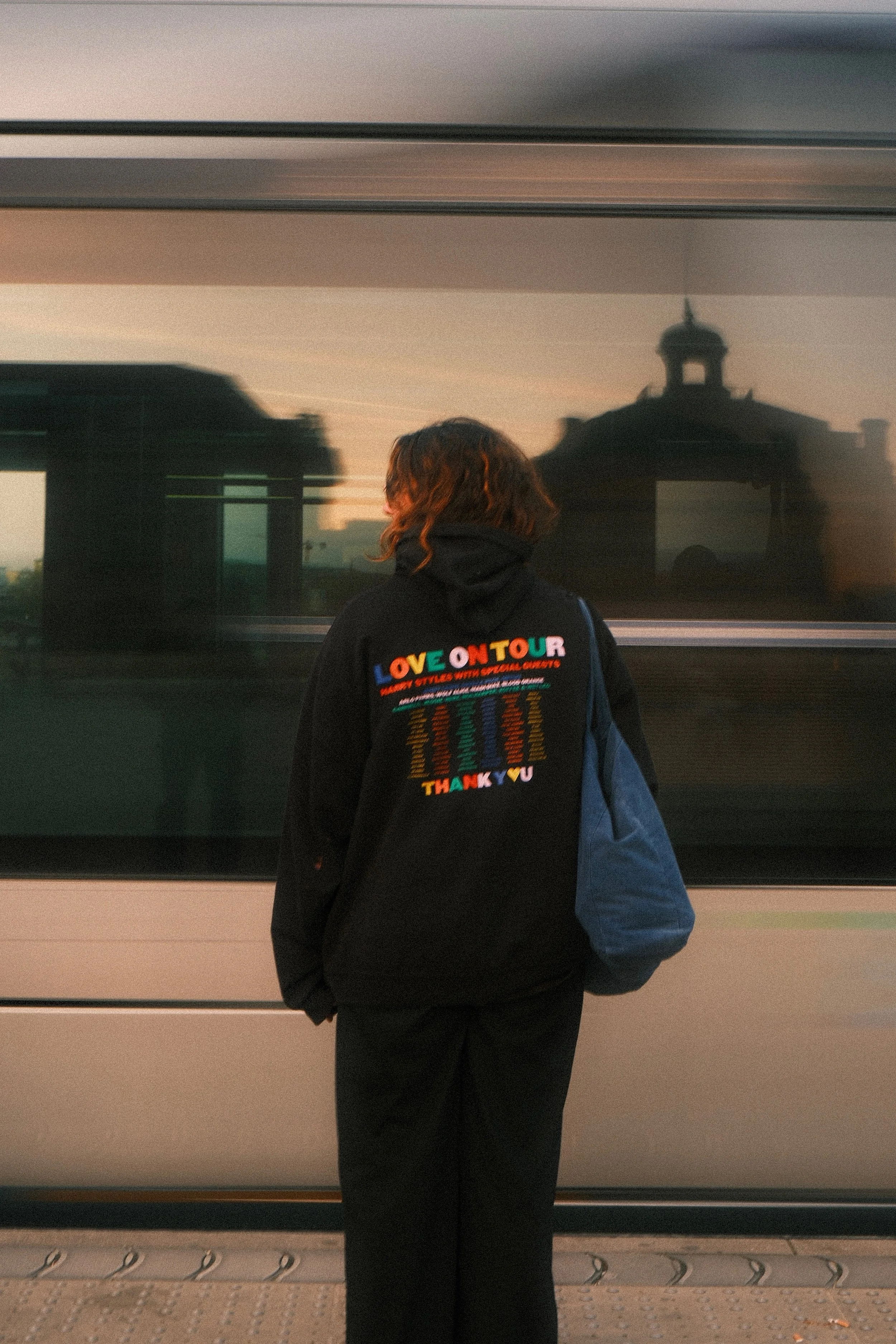 Une personne avec des cheveux roux, portant un sweat à capuche noir avec un message coloré à l'arrière, regarde un train en mouvement à l'extérieur lors du coucher du soleil.