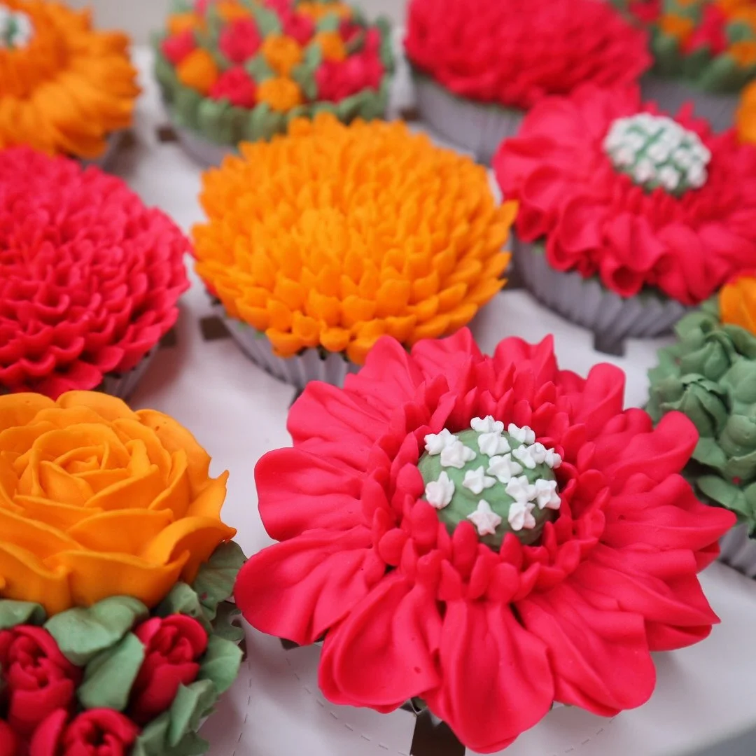 Colorful, intricately decorated cupcakes resembling flowers in various shades of pink, orange, and red with green accents, arranged on a white surface.
