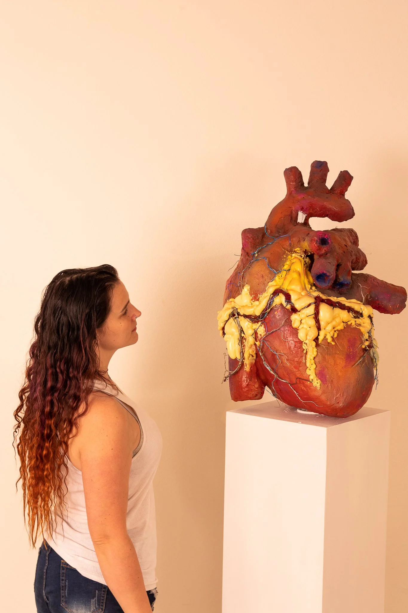 A woman with curly brown hair looks at a large, abstract sculpture of a human heart on a white pedestal in an art gallery.