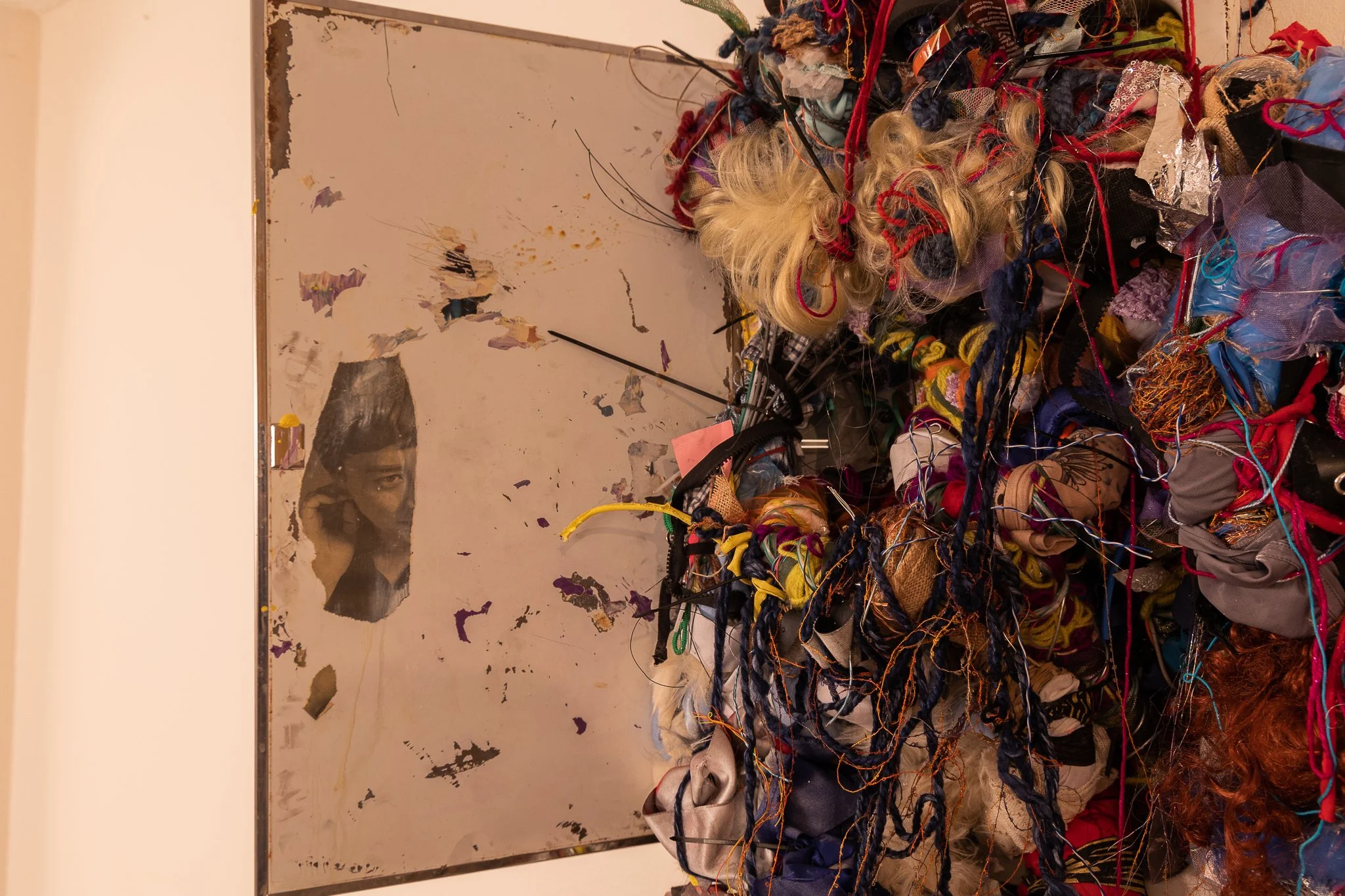An art installation featuring a pile of tangled multicolored yarn, fabric scraps, and wigs in front of a weathered mirror with torn paper and partial face reflection.
