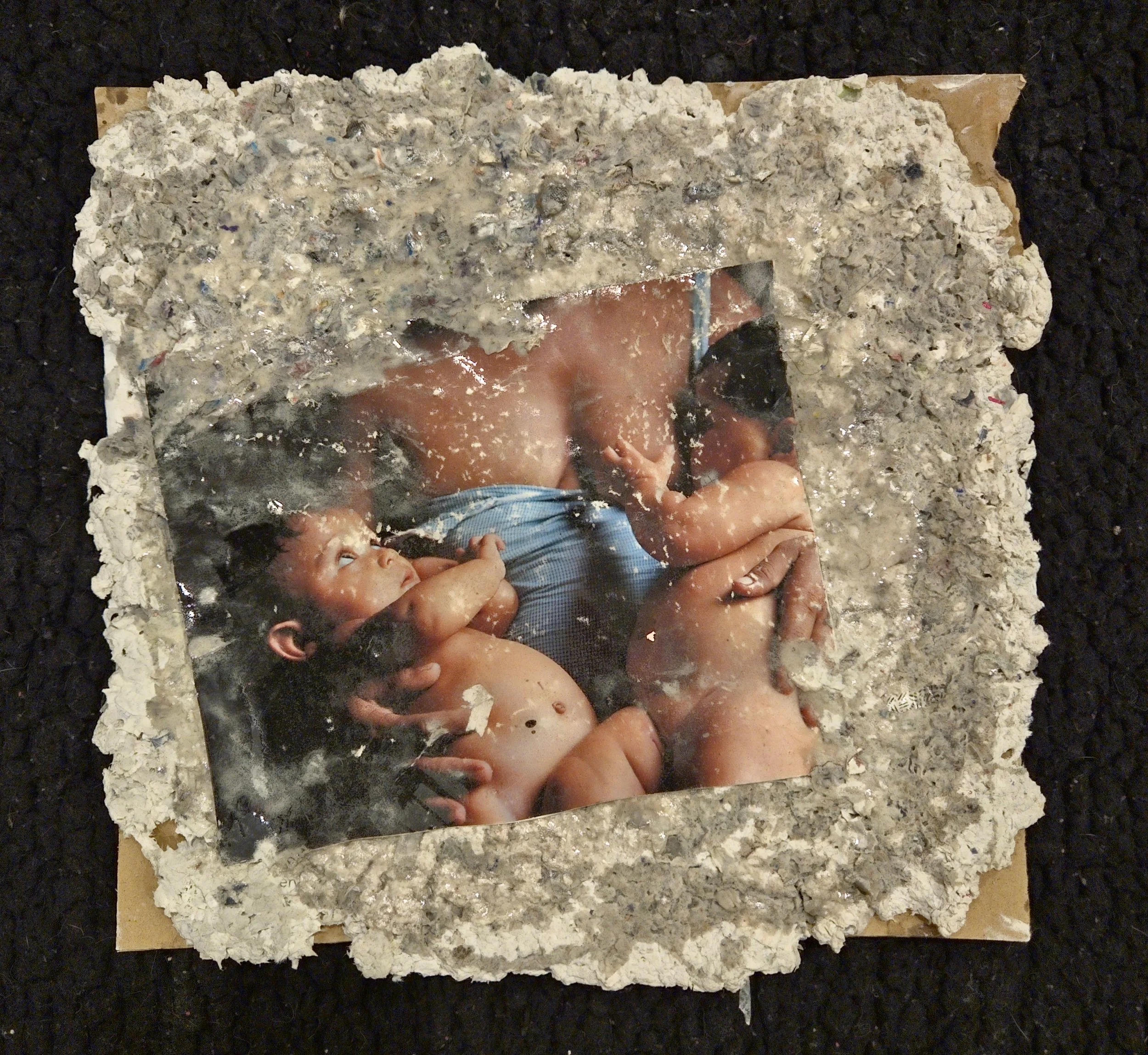 Framed photograph of a person holding a newborn baby, with the frame surrounded by a rough, textured material resembling concrete or cement, and the photo appears to have some damage or wear.