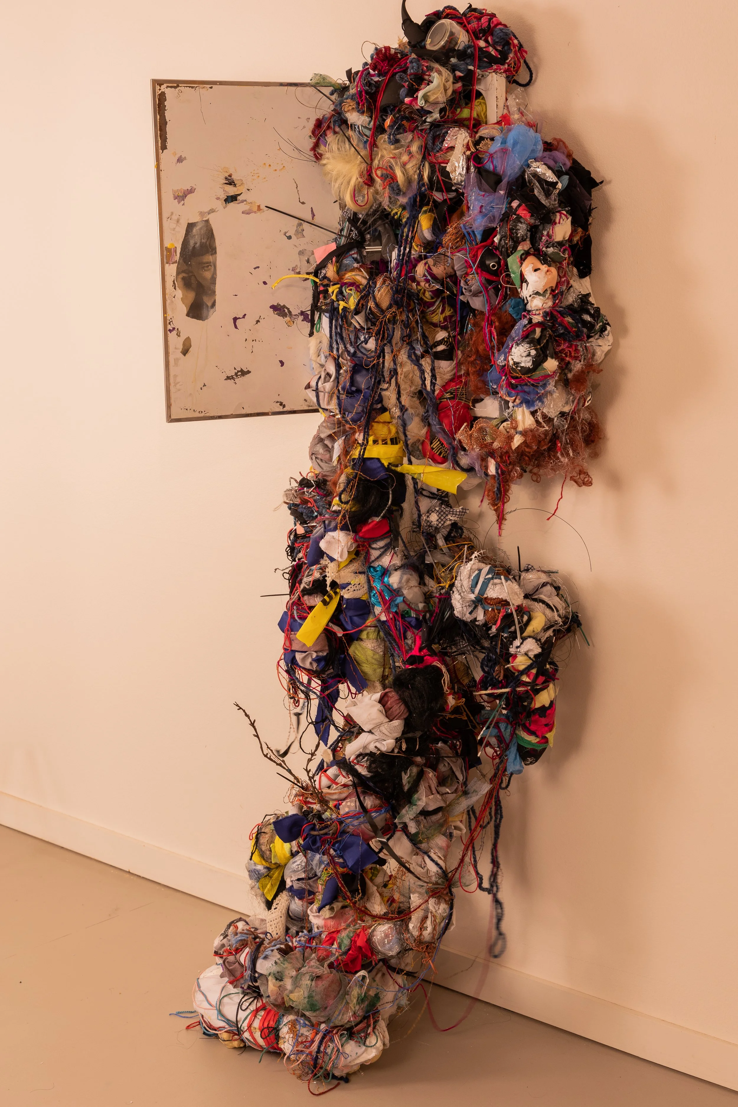 A large sculpture made of tangled colorful fabric, yarn, and miscellaneous materials, leaning against a white wall. A framed art piece with a partially visible face is in the background on the wall.