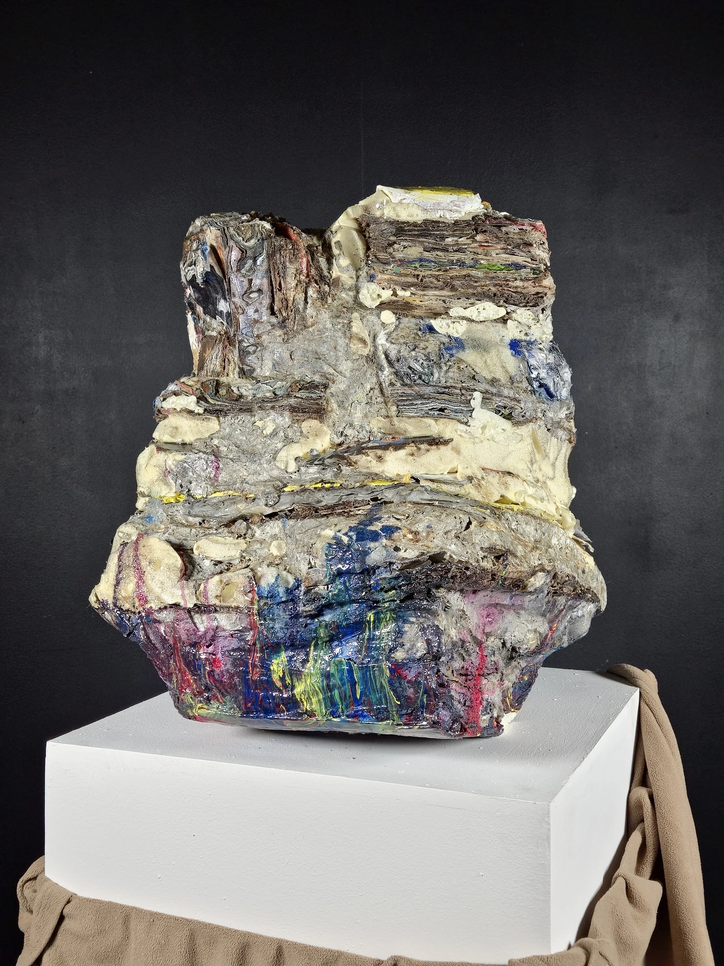 Mixed media sculpture resembling a humorous face, placed on a white pedestal against a dark background.