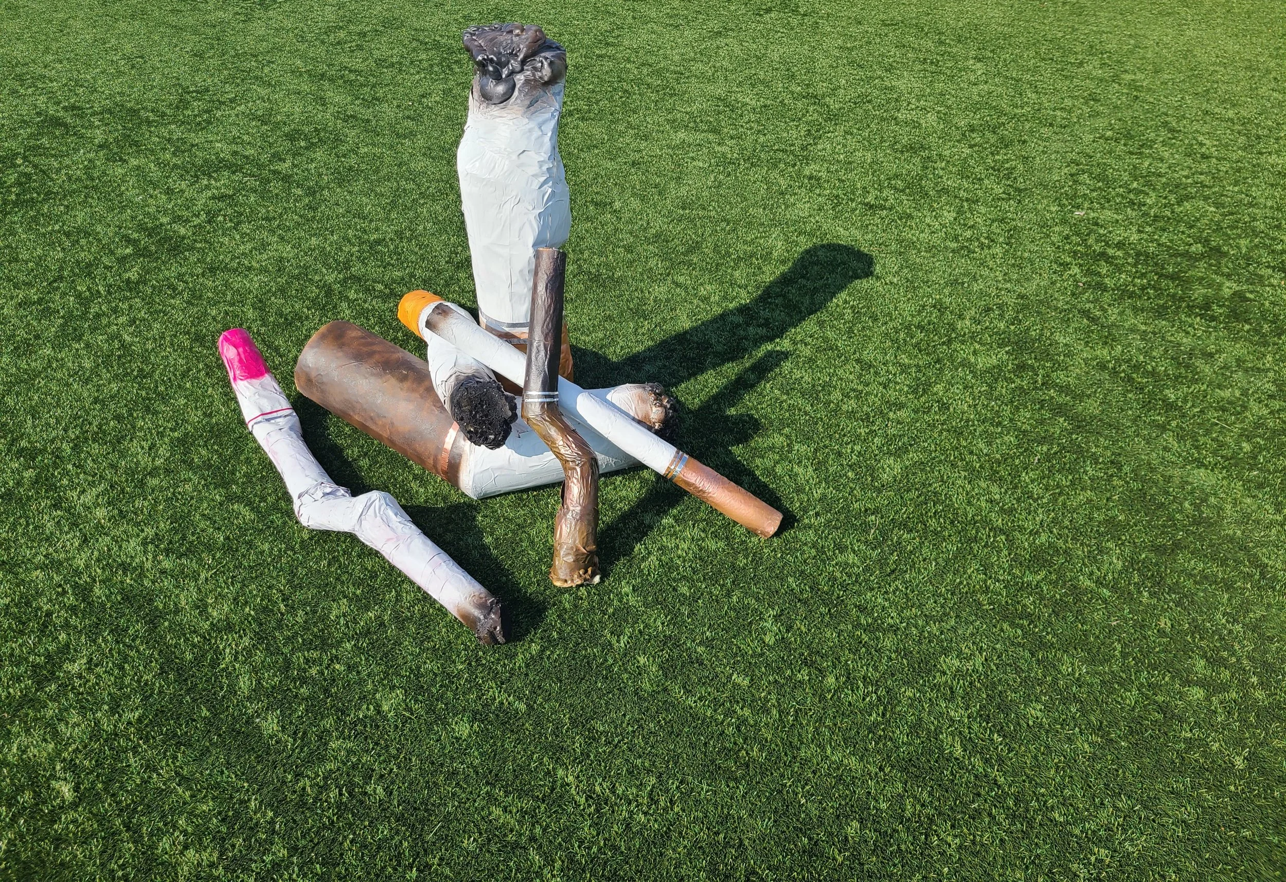 Colorful sculpture of a sitting cat made from painted and wrapped materials, placed on a grassy field, with its shadow cast to the right.