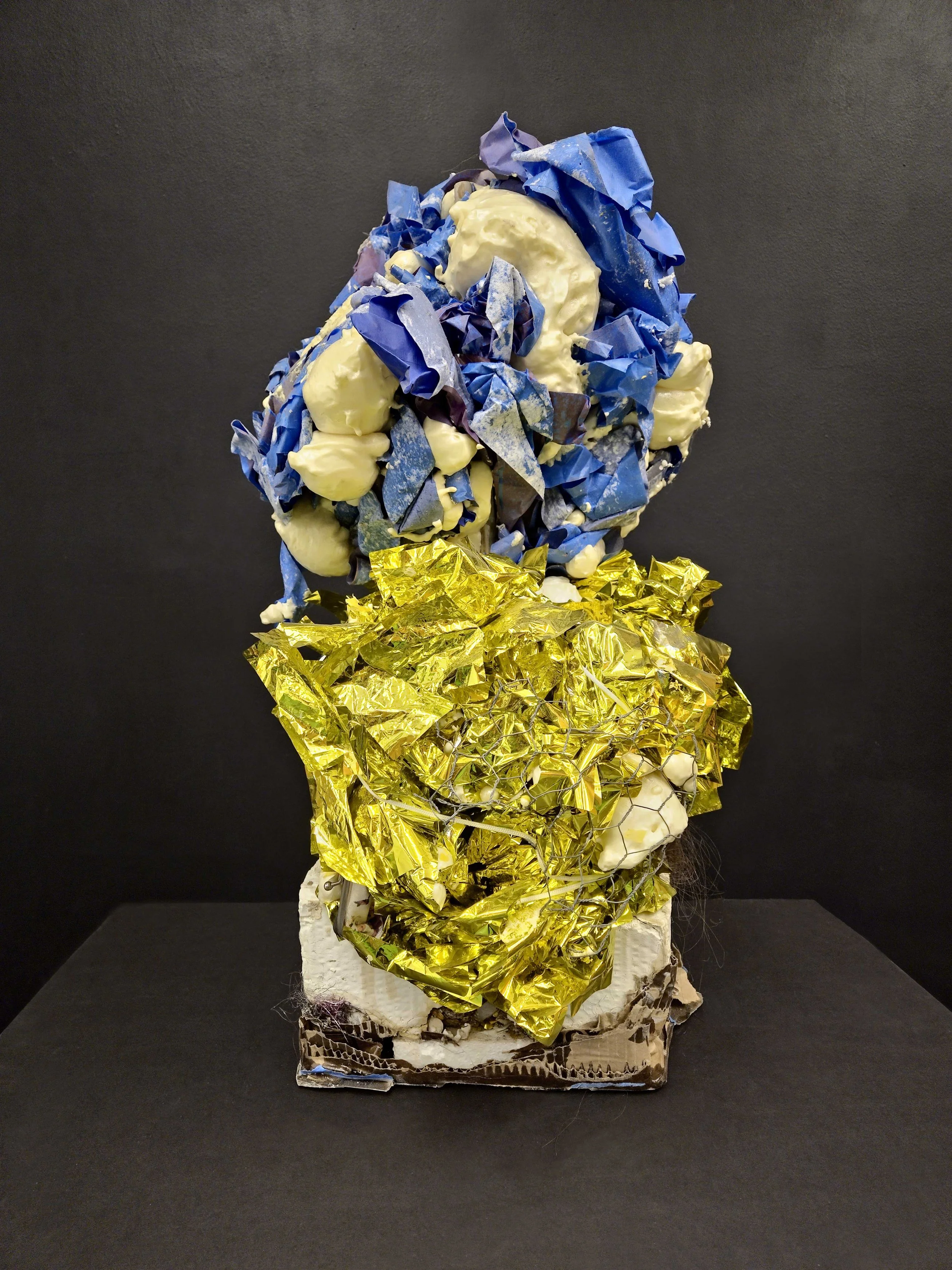 Mixed media sculpture with a black background, featuring a top section of crumpled blue and white paper, a middle section of yellow crumpled metallic foil, and a base made of layered materials.