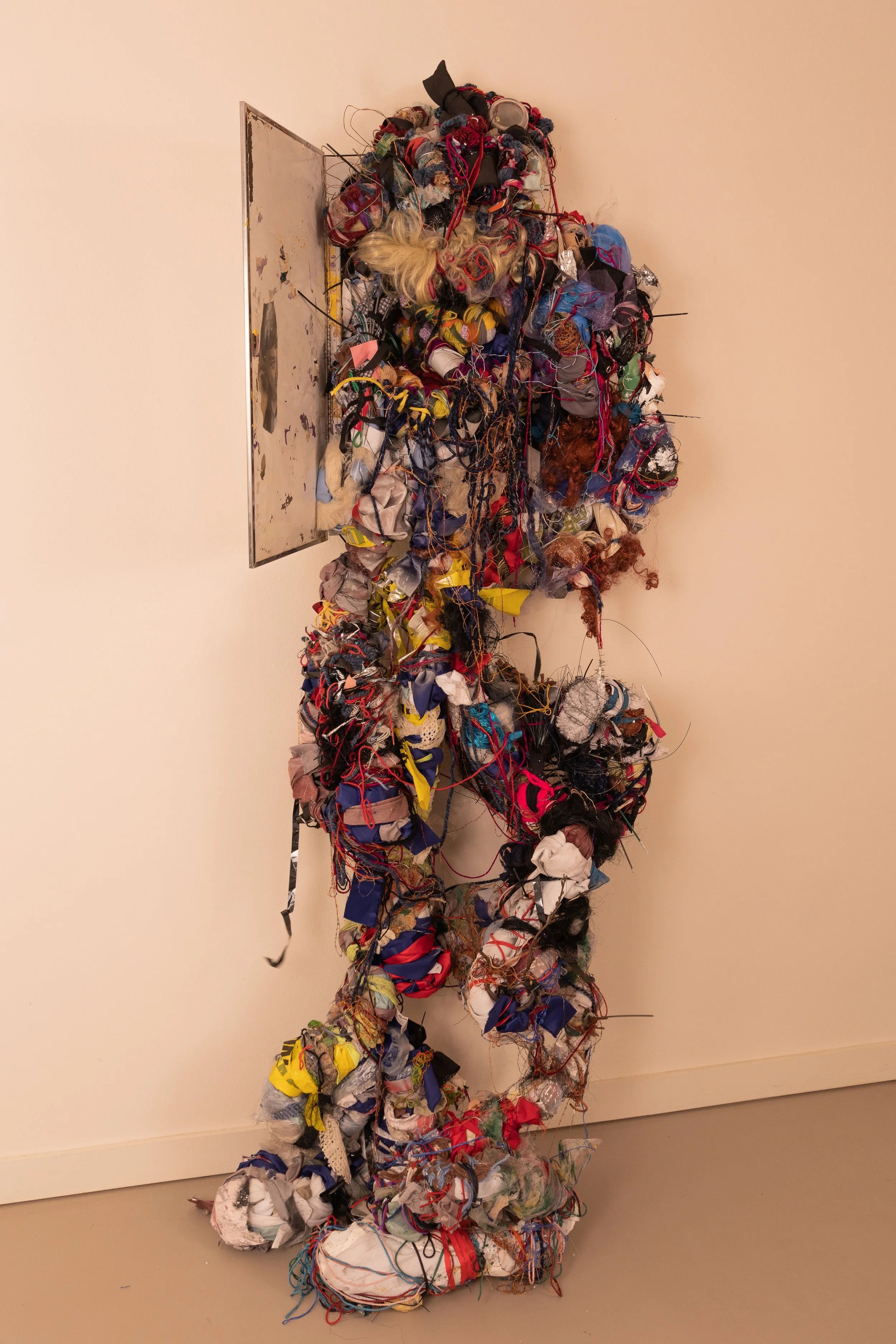 Messy art installation made of tangled yarn, fabric scraps, and assorted textiles, with a metal sheet attached to the wall behind it.