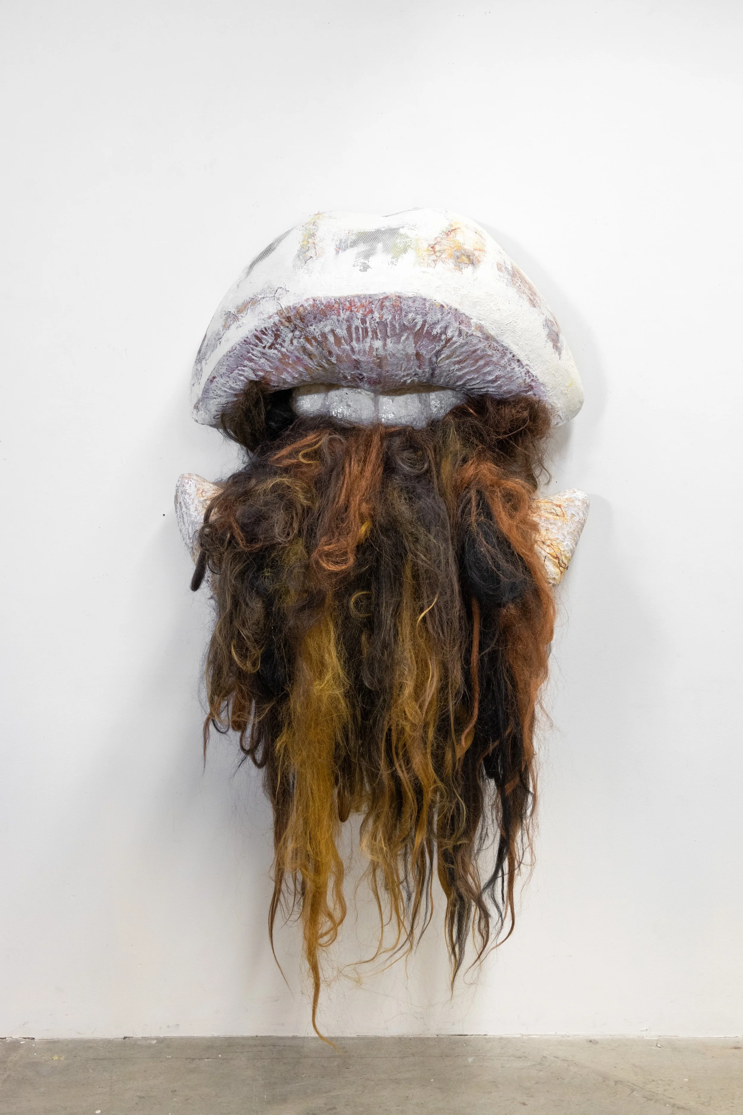 Art sculpture of a person's face with large lips and a bushy beard made of mixed-colored hair, mounted on a white wall.