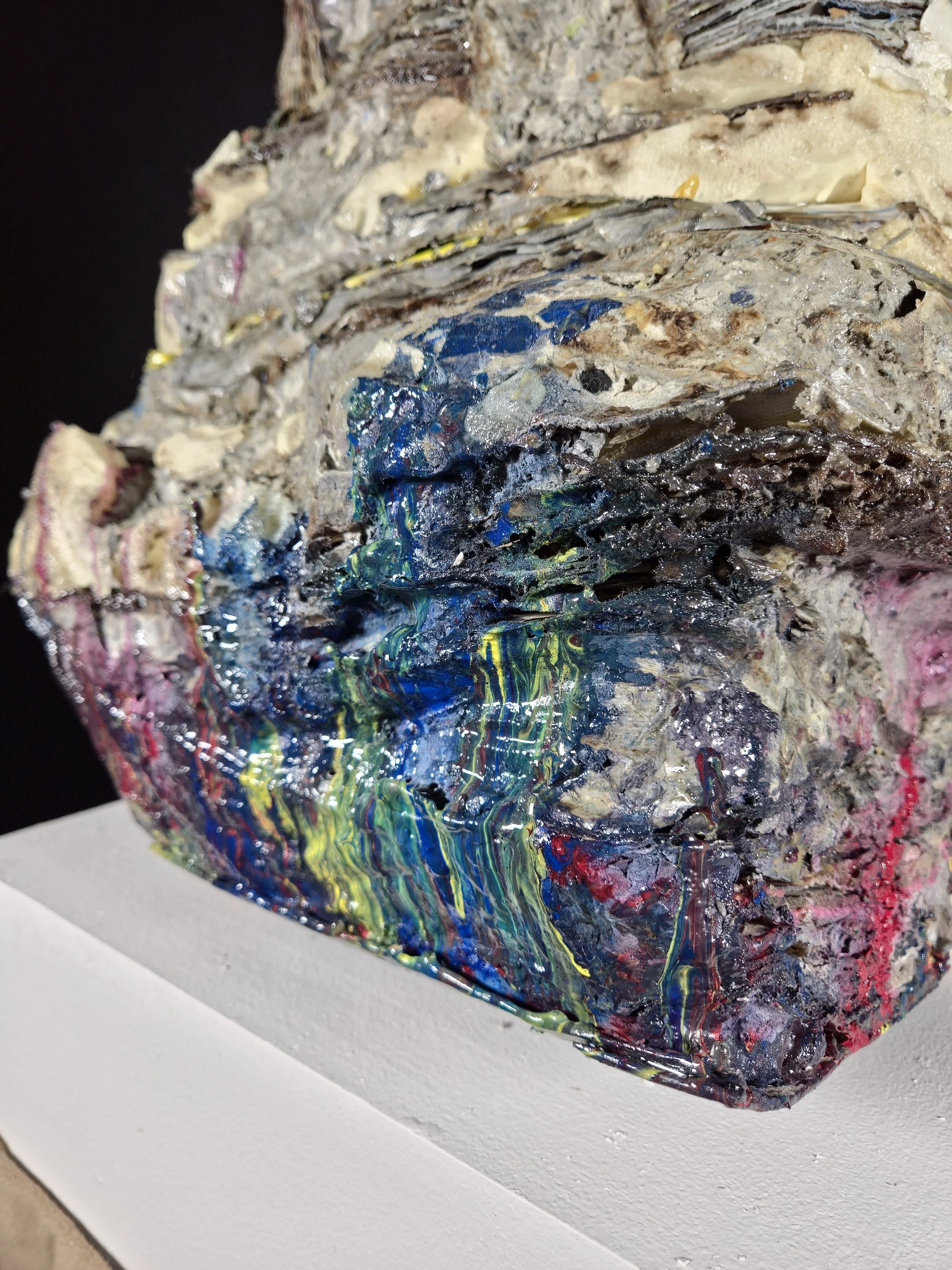 A textured sculpture with layered, rough material in neutral tones mixed with vibrant, glossy streaks of blue, green, red, yellow, and black, placed on a white pedestal.
