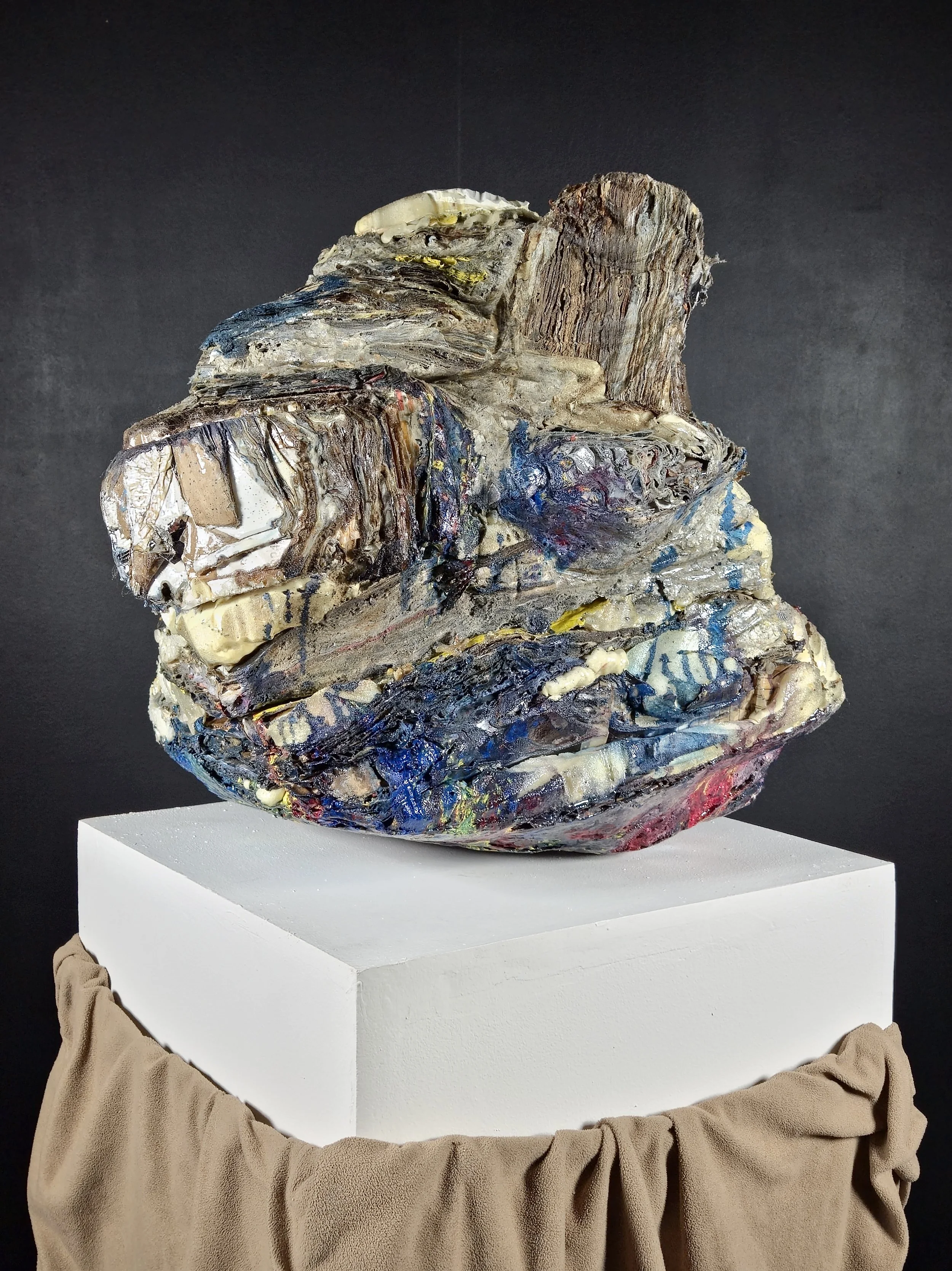 A textured sculpture resembling a human face made from mixed media, placed on a white platform with a brown fabric draped around its base, set against a black background.