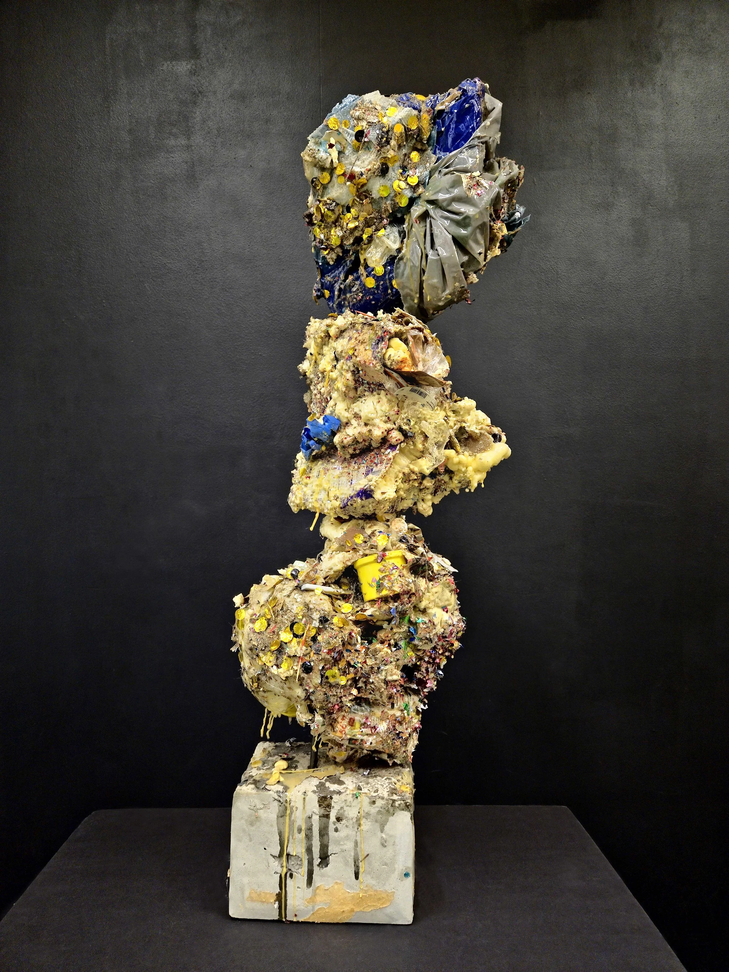 Abstract sculpture made of trash and debris, stacked vertically on a concrete base, against a black background.