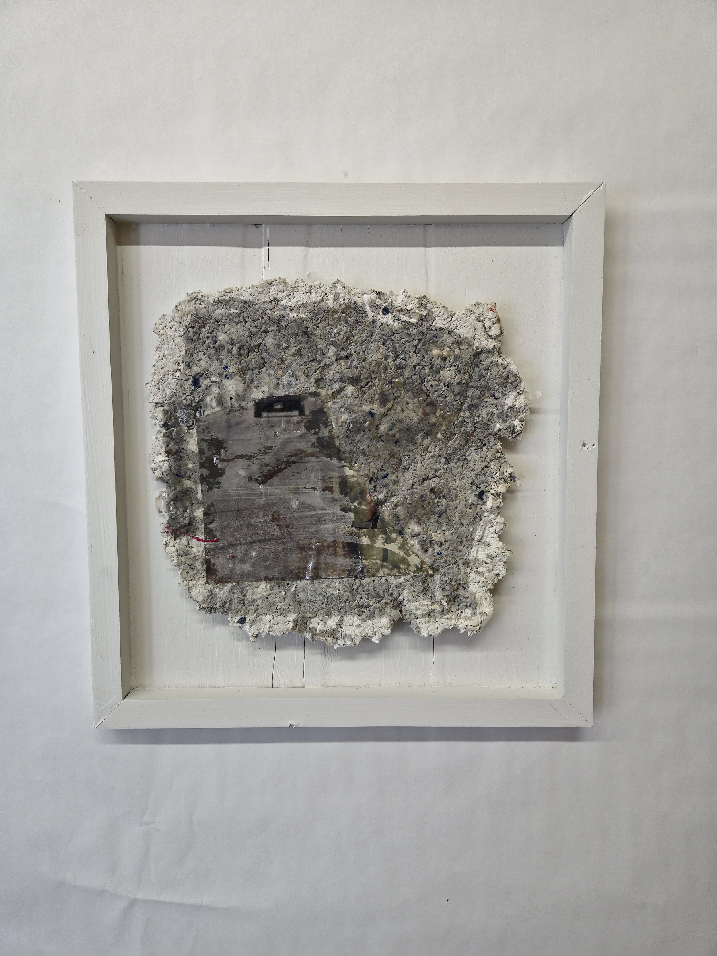 Mixed media artwork mounted in a white shadow box frame, consisting of rough textured gray material with embedded transparent sections.