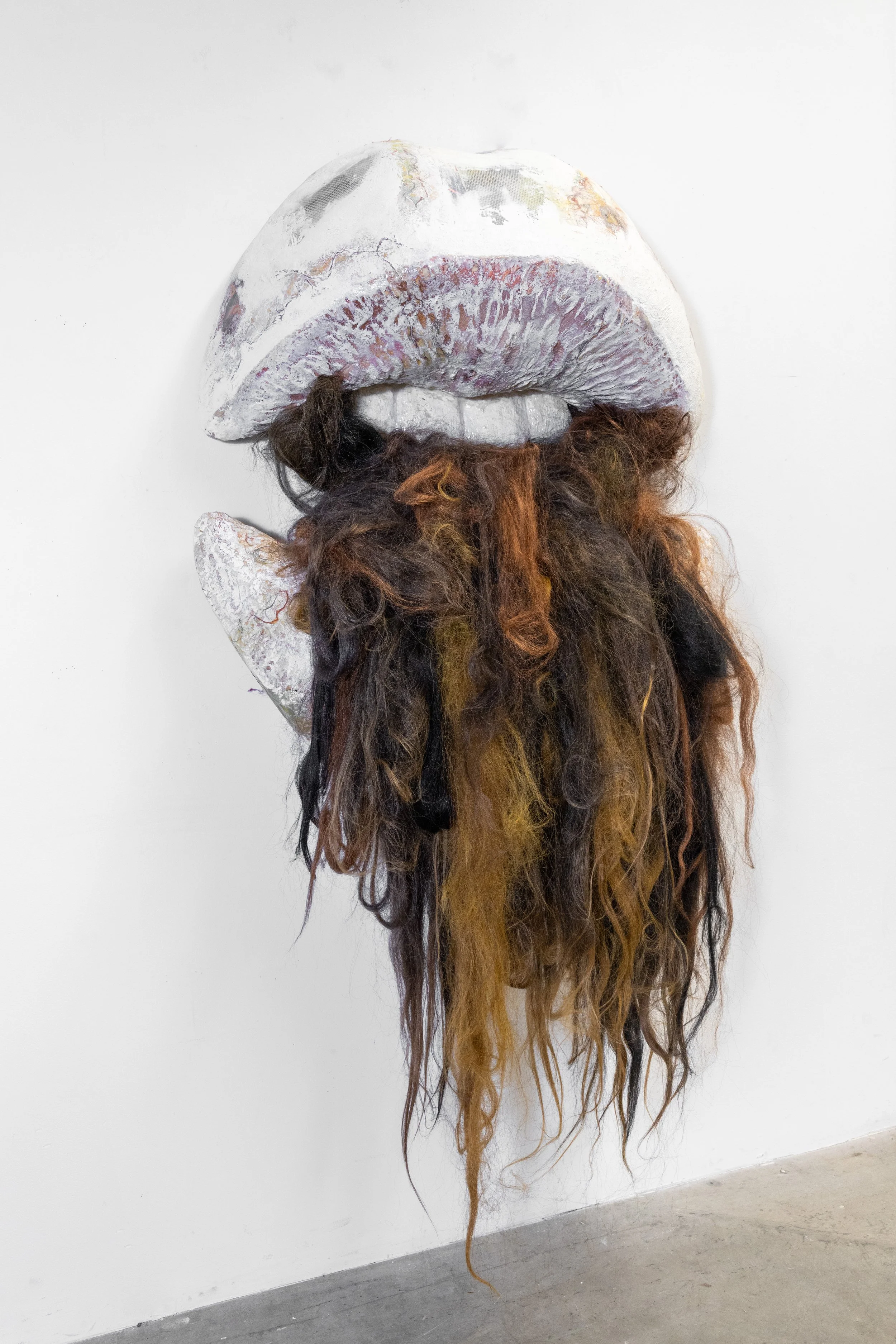 Mixed media art piece resembling a face with large lips and a long beard, with lips made of clay and hair-like materials for the beard, mounted on a white wall.