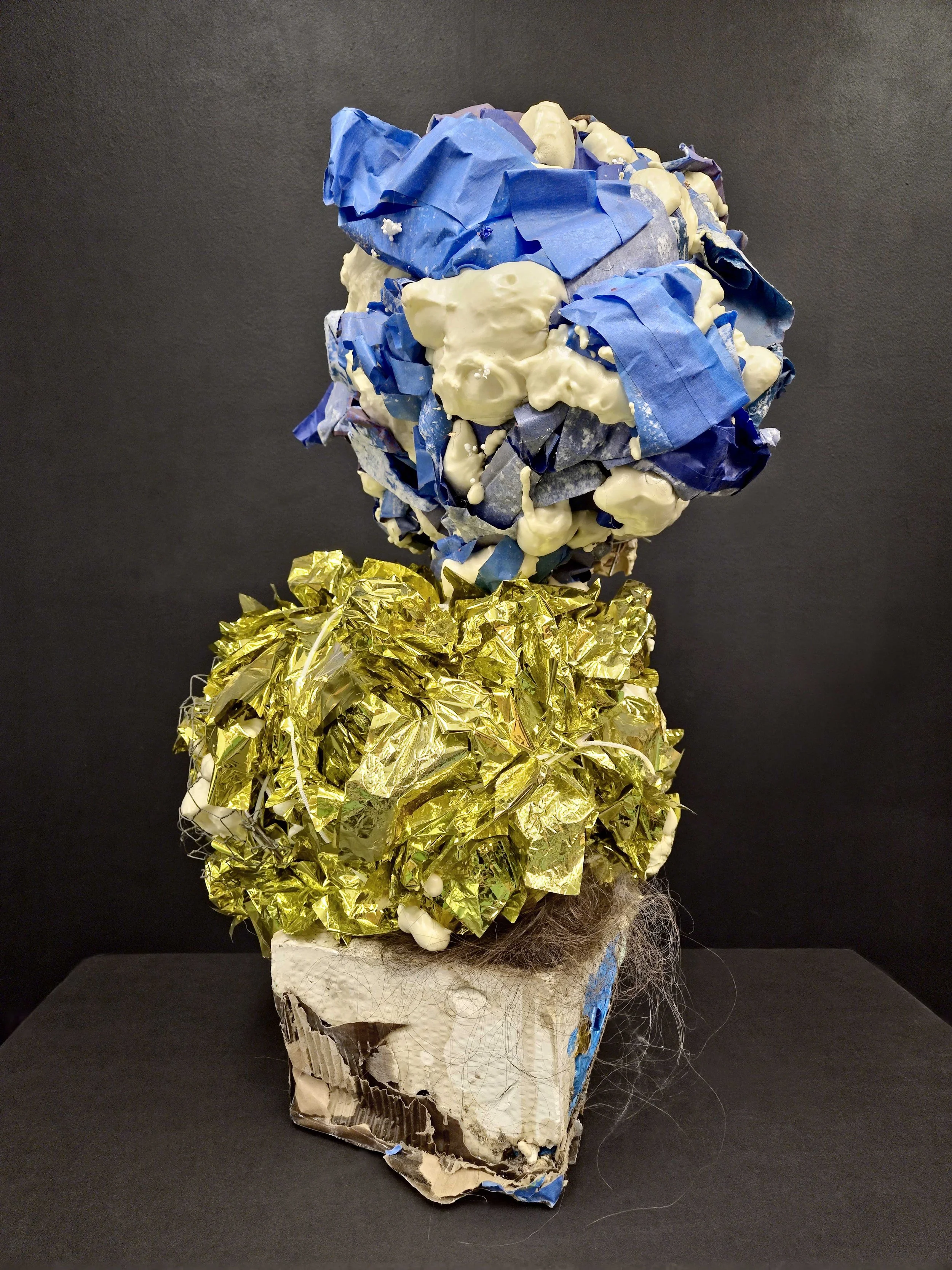 Mixed media sculpture resembling a flower with a dark background, made from crumpled paper, foil, fabric, and synthetic fibers.