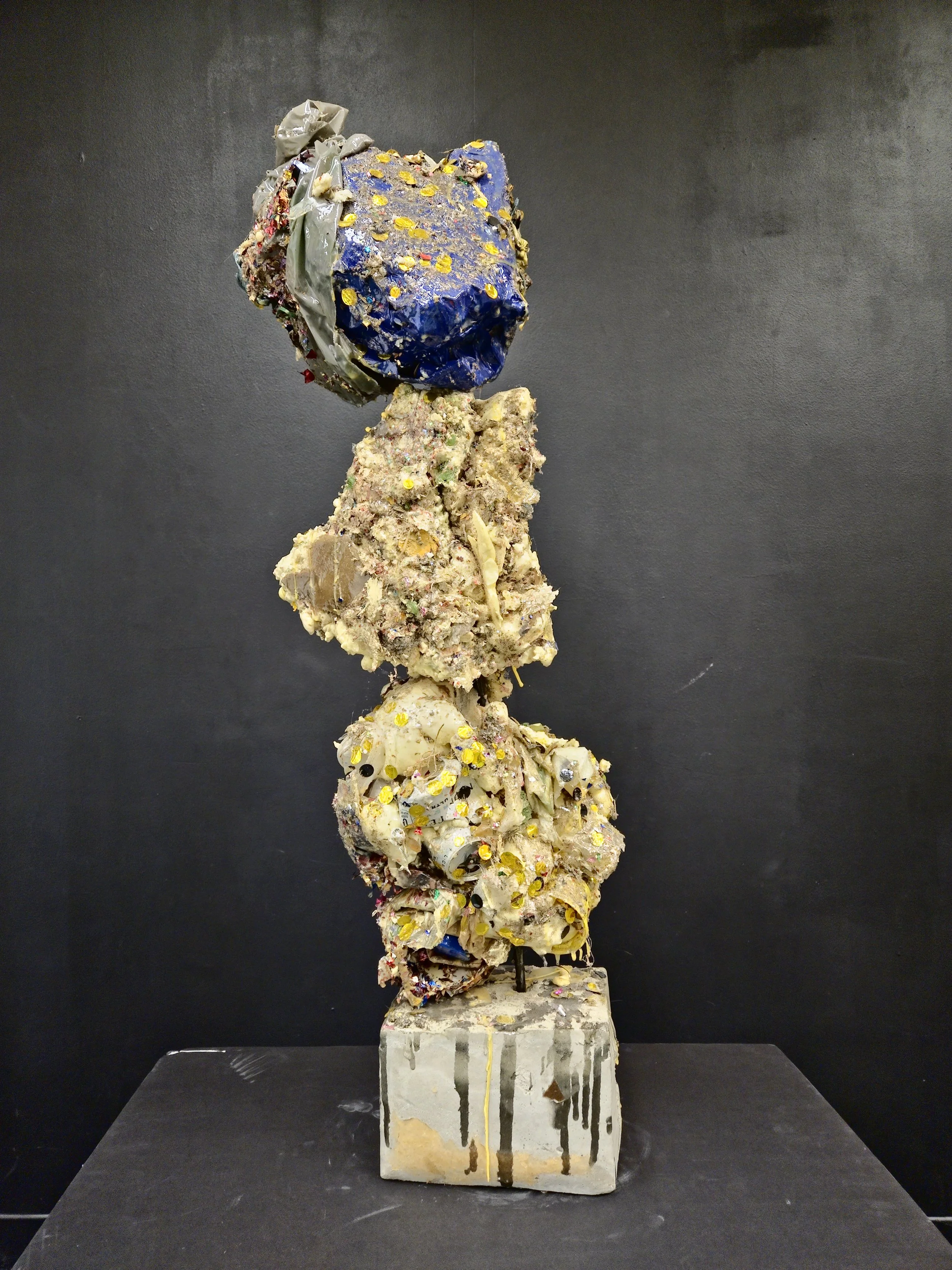 Mixed media abstract sculpture with various textured and colorful materials stacked on a concrete base against a black background.