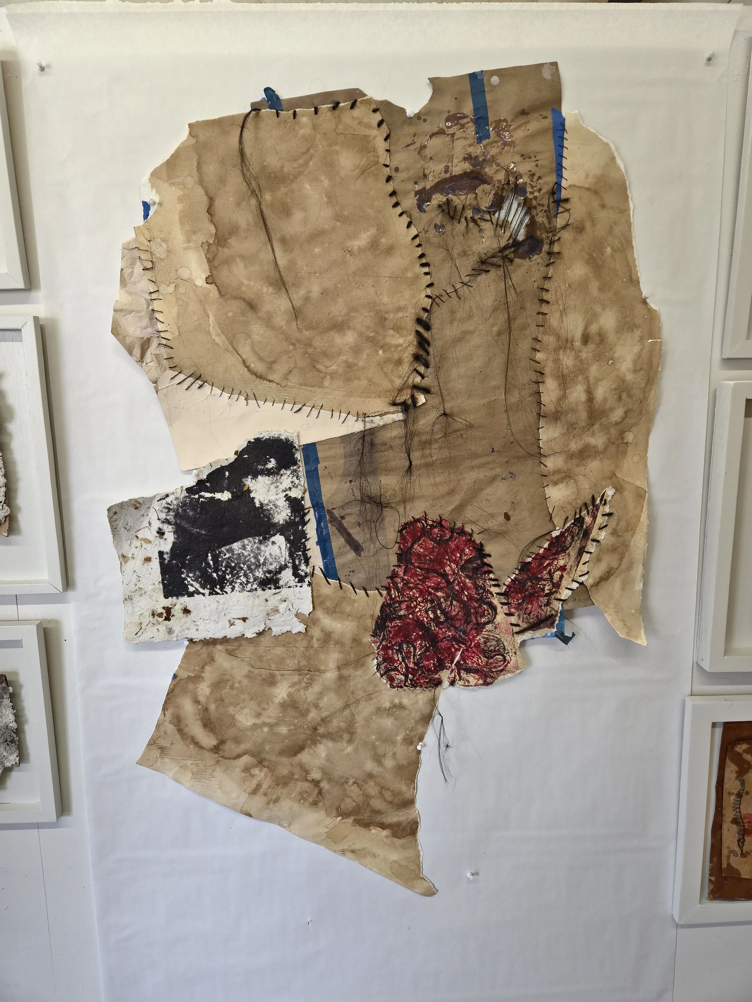 Mixed media abstract artwork on torn paper with stitched outlines, water stains, black and white paint, and red textured patches, hanging on a white wall.