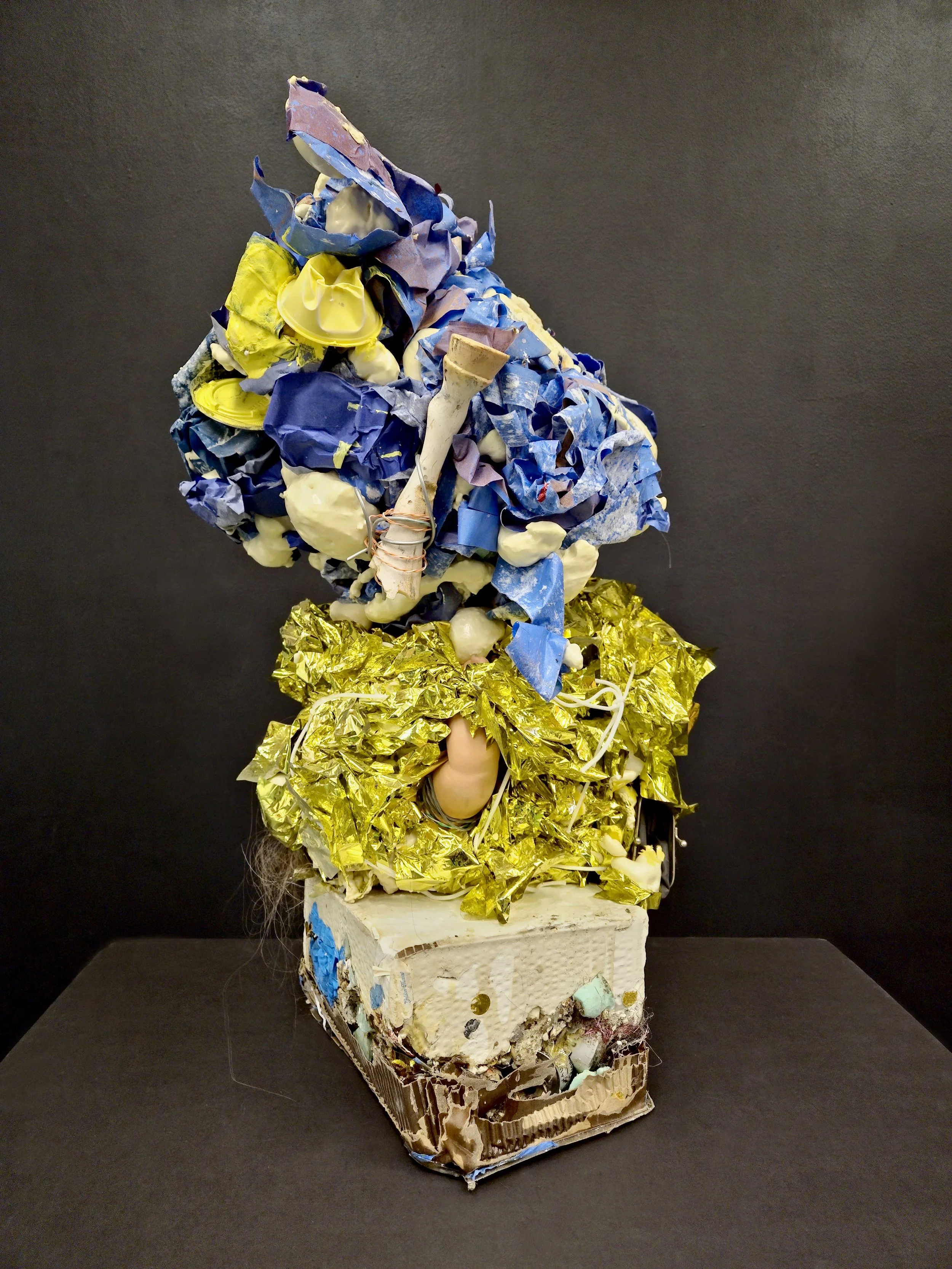 An abstract sculpture made from various crumpled and layered plastic materials, including blue, yellow, and gold-colored items, arranged on a white base with a dark background.