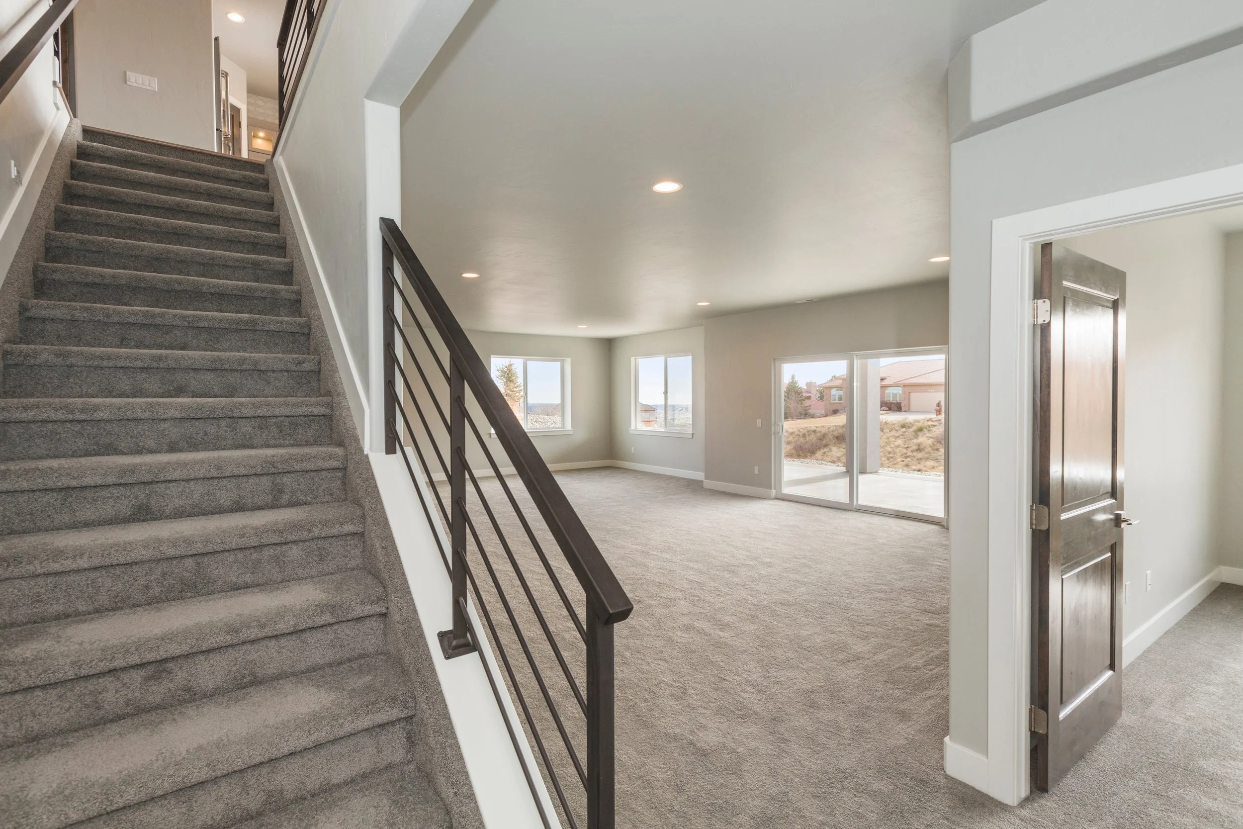 Interior of a modern home with gray carpeted stairs, black metal handrail, and spacious living area with large windows and sliding glass doors leading outside.