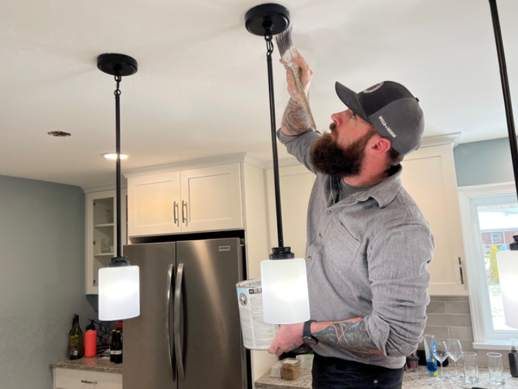 A man with a beard, tattoos, and wearing a cap, painting the ceiling of a kitchen with a paint roller.