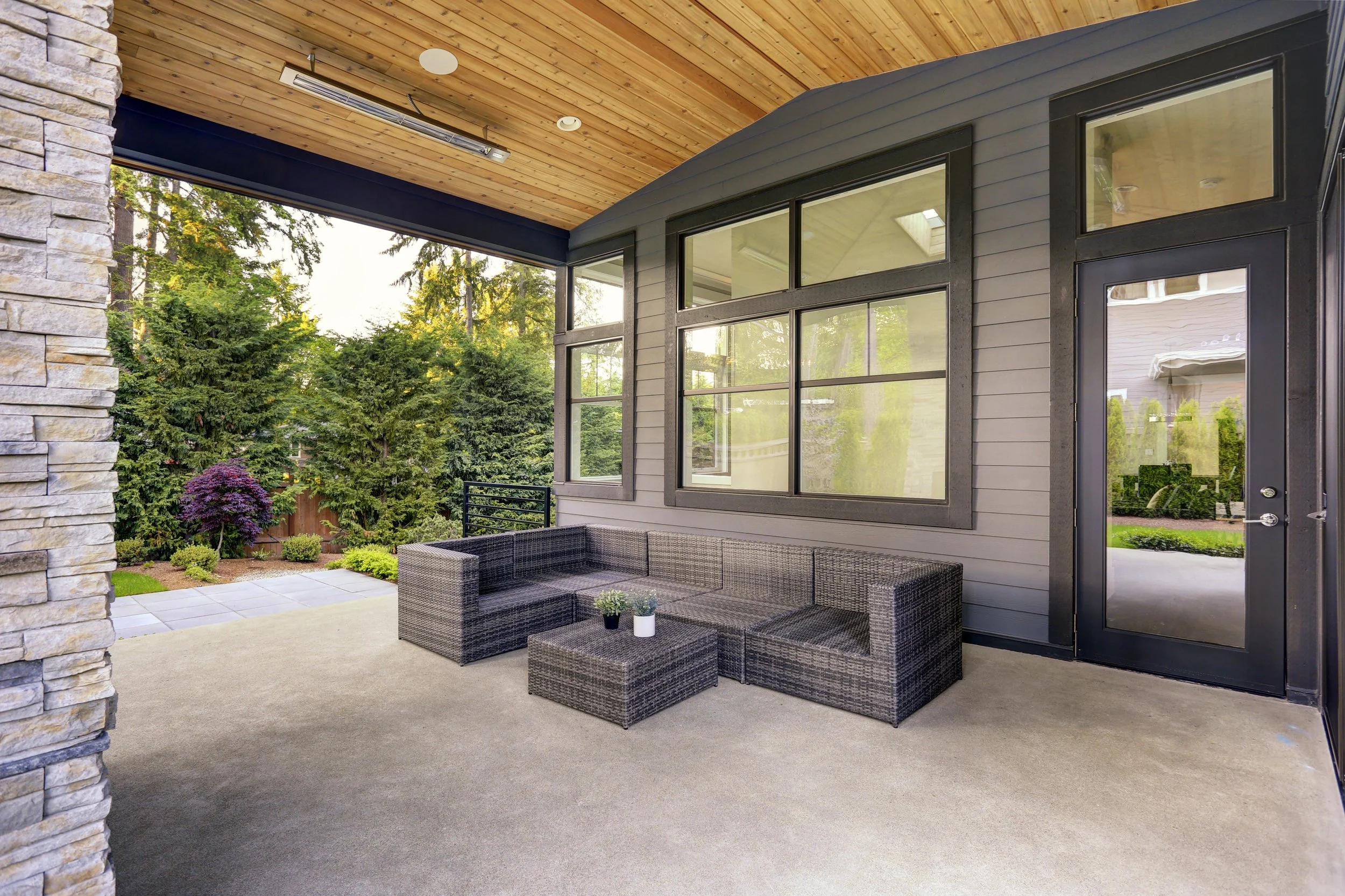 Outdoor patio with wicker furniture, a stone wall, large windows, and lush greenery