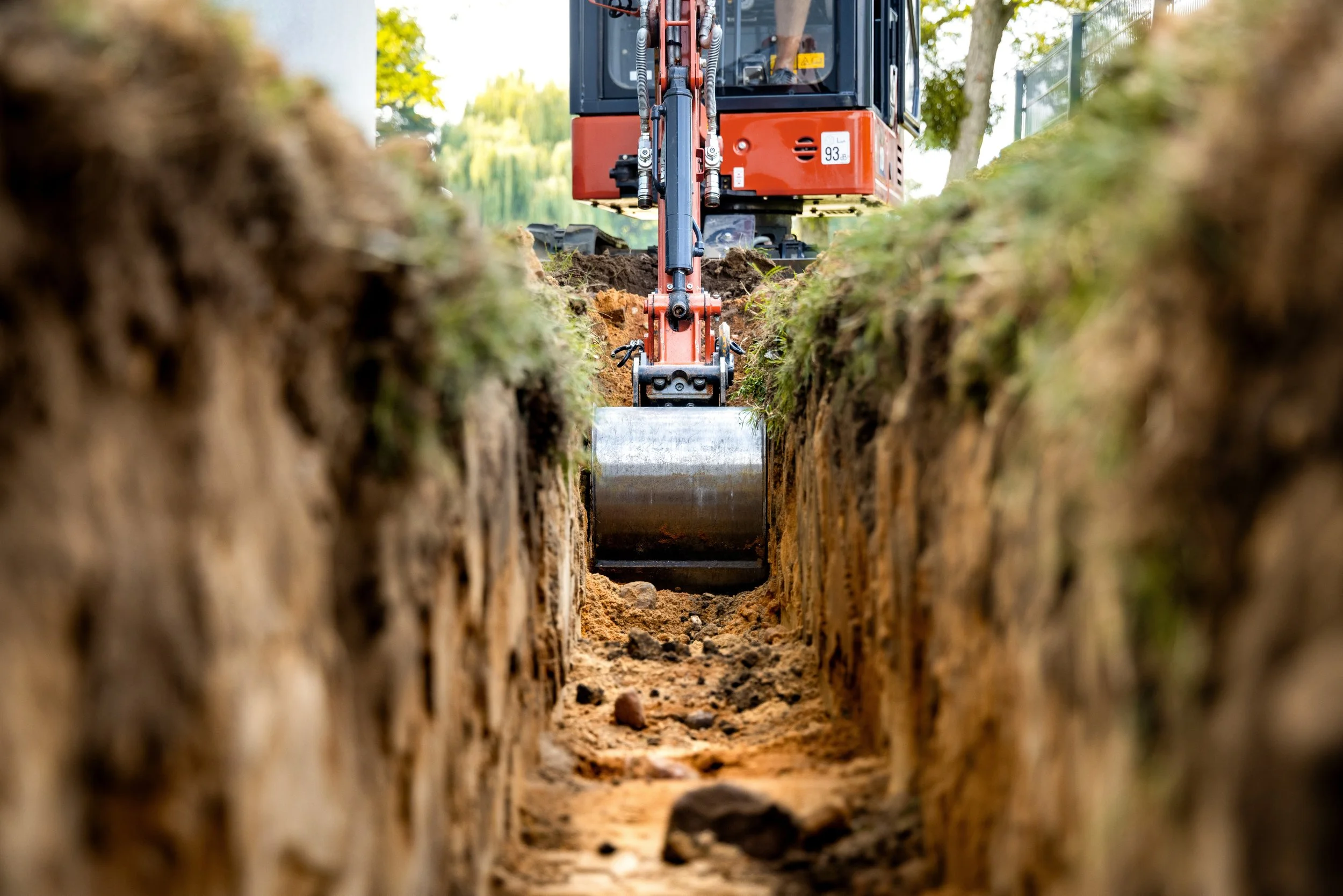 Close-up view of a construction excavator working in a trench, with the camera positioned between the walls of the trench, focusing on the excavator's arm and bucket.