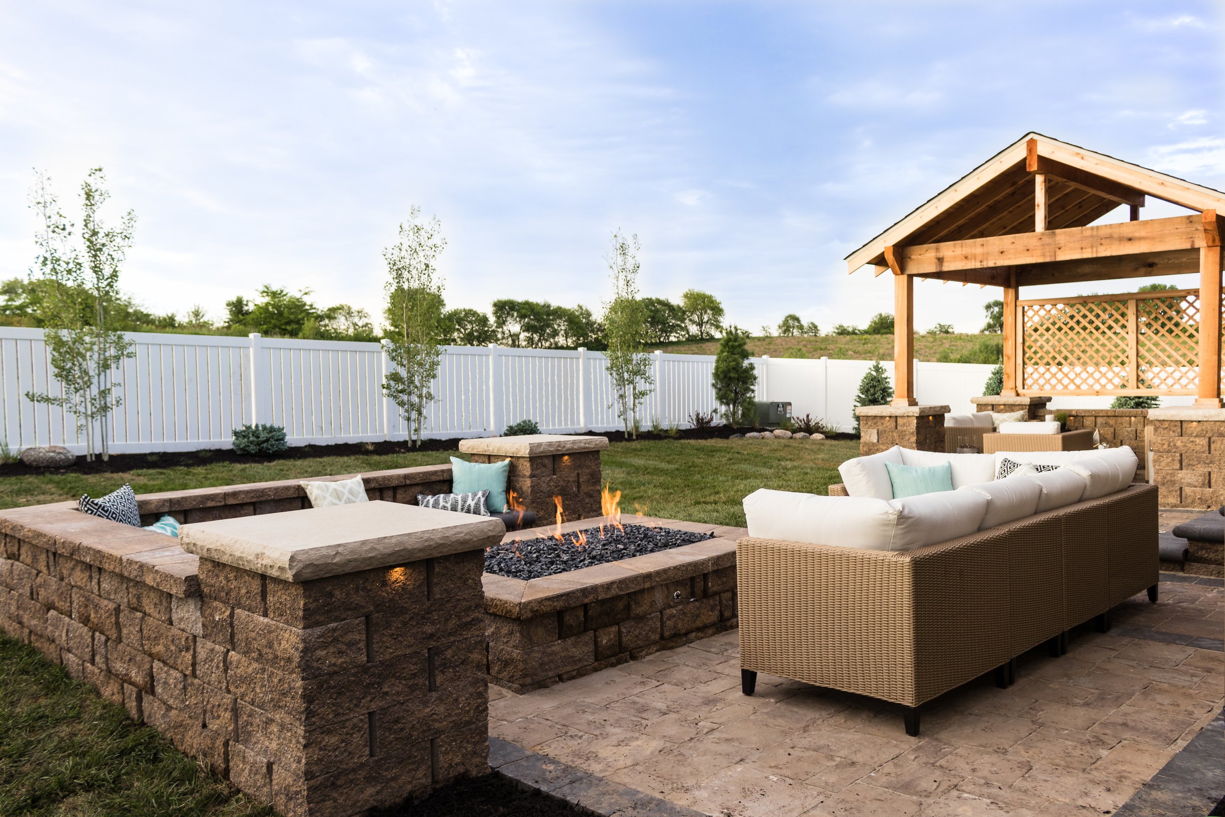 Backyard patio area with seating, a fire pit, and a wooden pergola, enclosed by a white fence, with trees and a cloudy sky in the background.