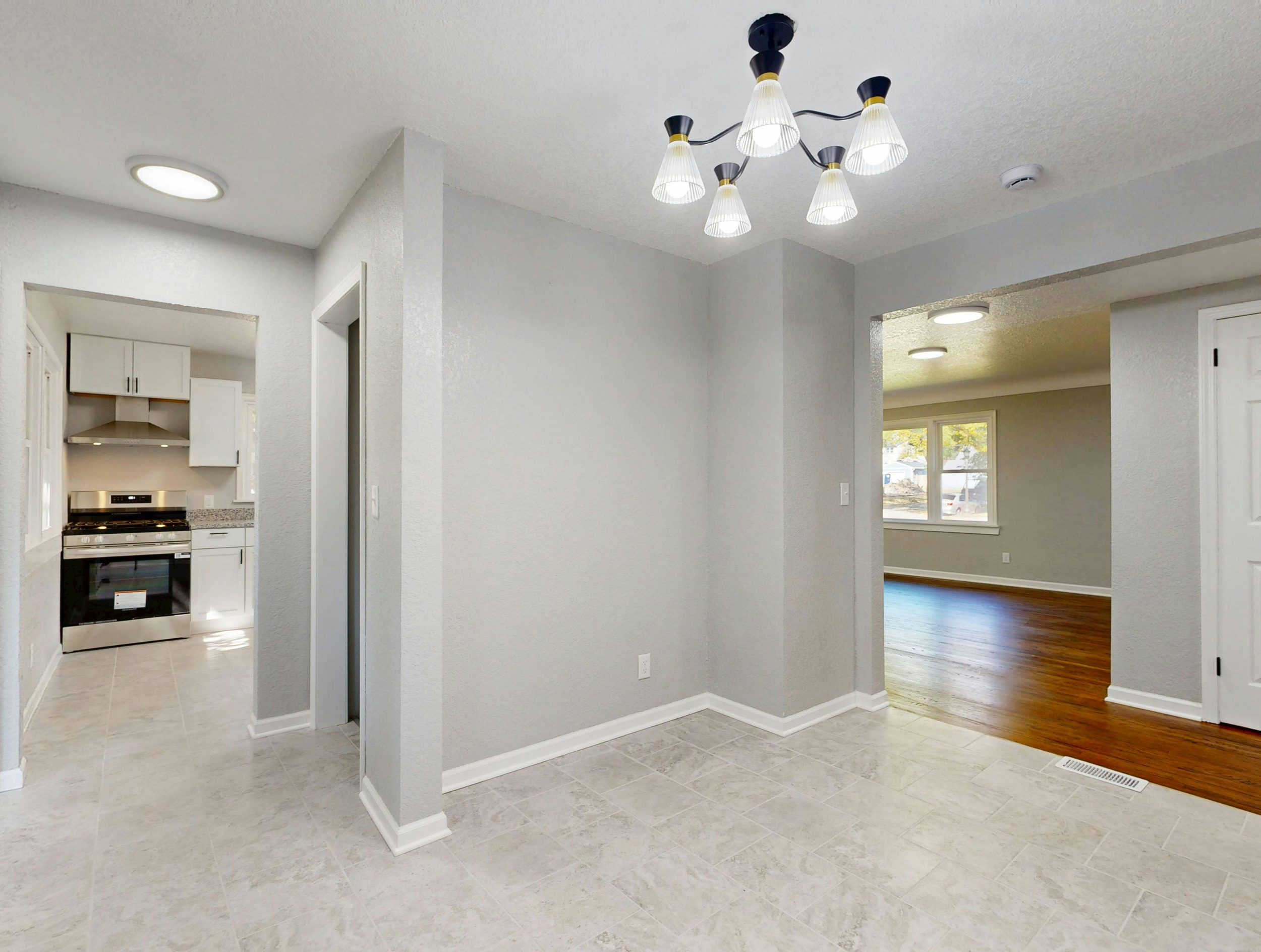 Interior view of a modern home with white walls, beige tiled flooring, a kitchen with white cabinets and a stainless steel oven, and a living area with wooden flooring, large windows, and multiple ceiling lights.