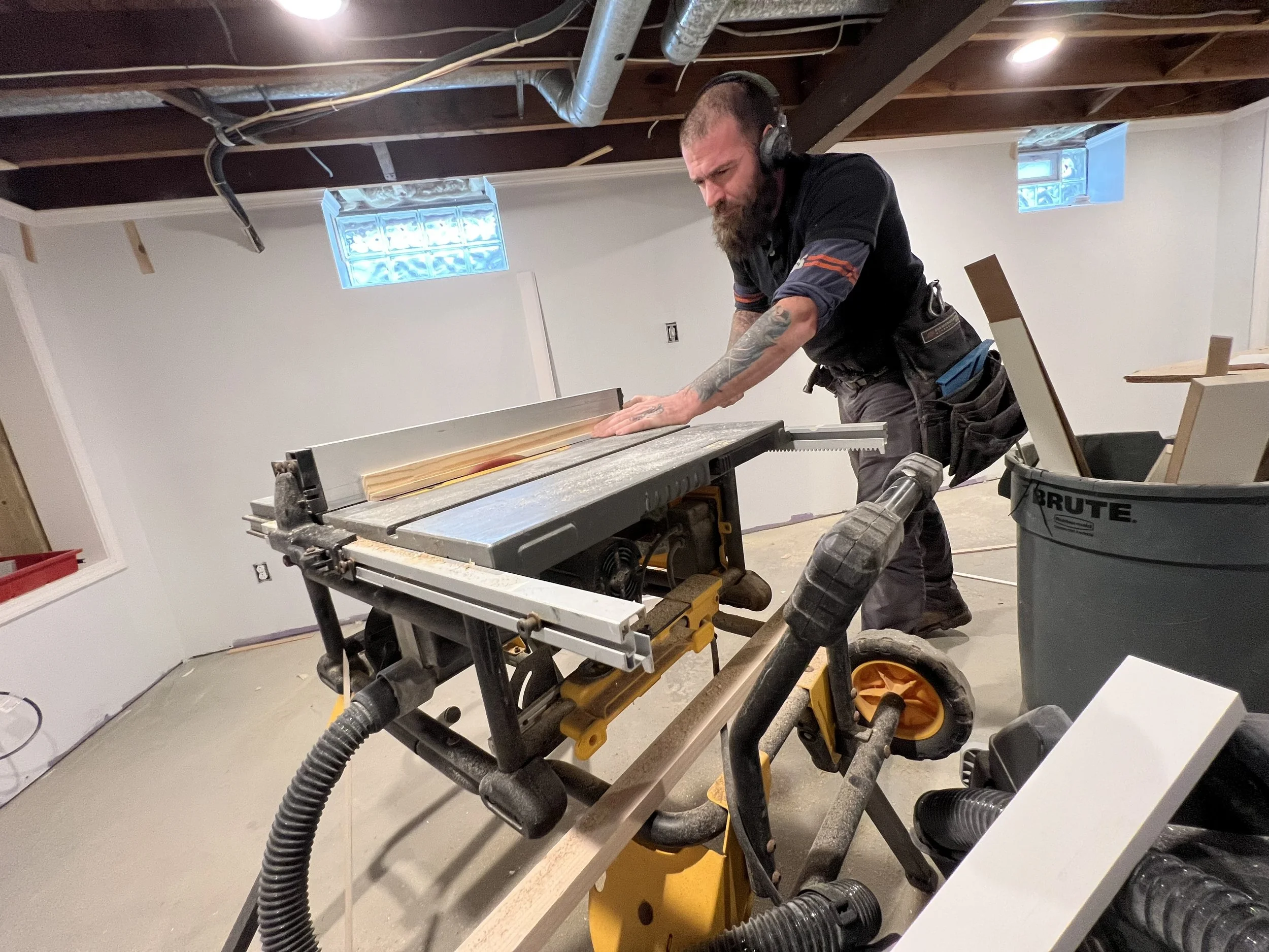 Man with tattoos wearing headphones operating a table saw in a basement renovation or construction site.